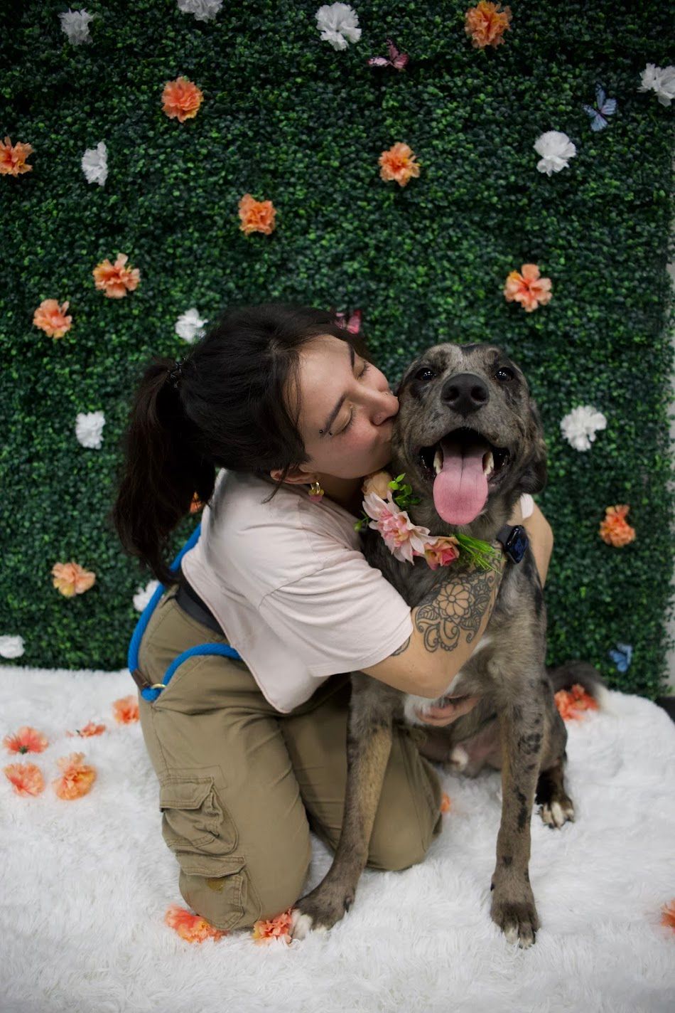 Woman kissing a gray dog wearing a floral collar in front of a green flower wall backdrop.