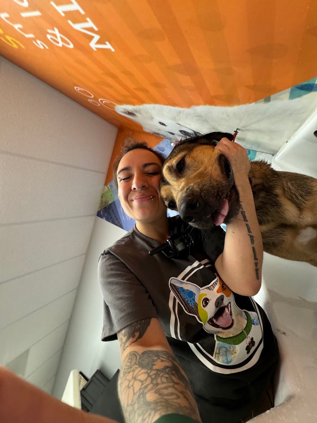 A smiling woman with tattoos gets a kiss from a dog while taking a selfie outdoors.