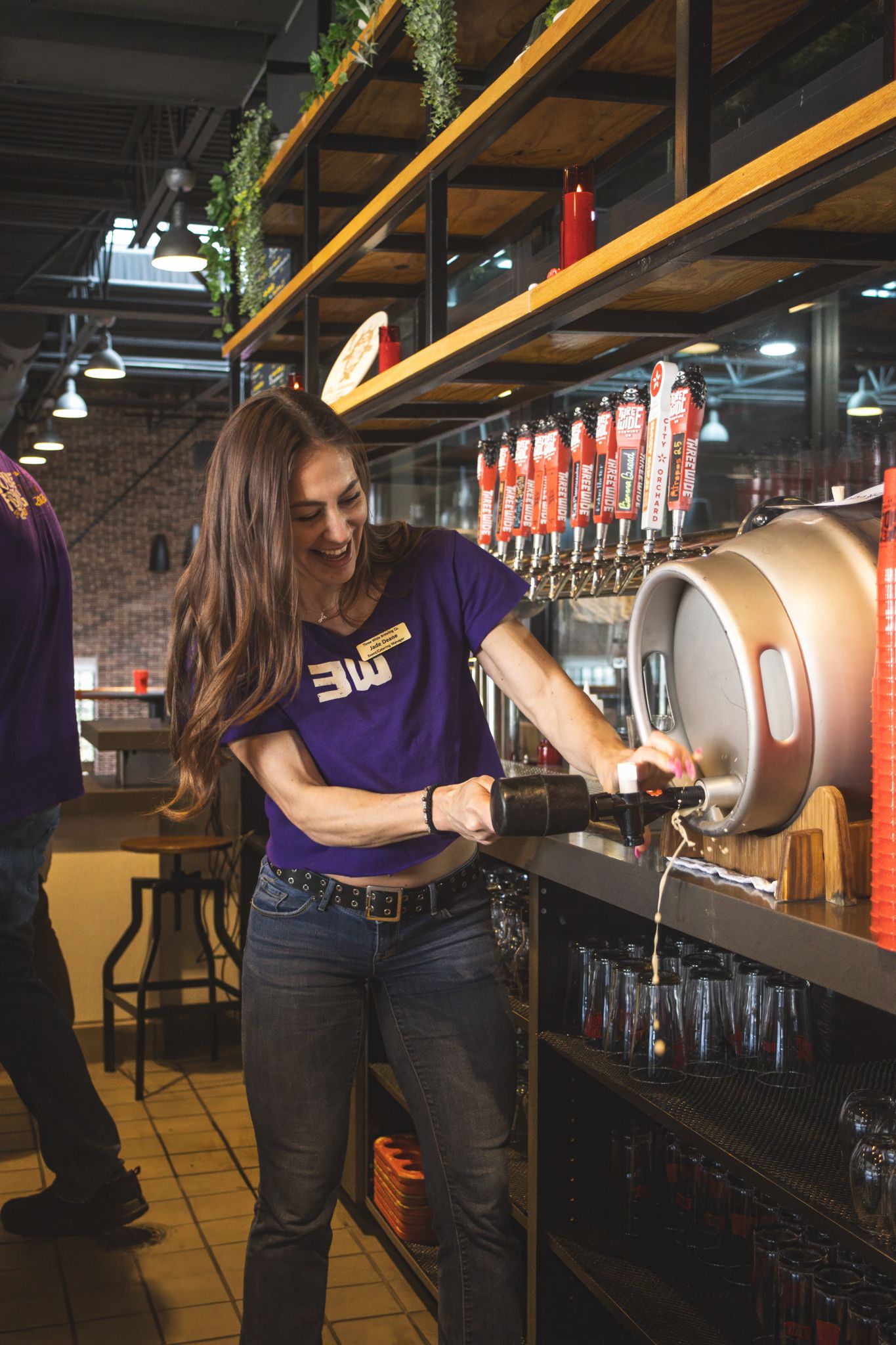 A smiling female bartender in a purple 3W shirt pours a drink from tap handles at a brewery bar.