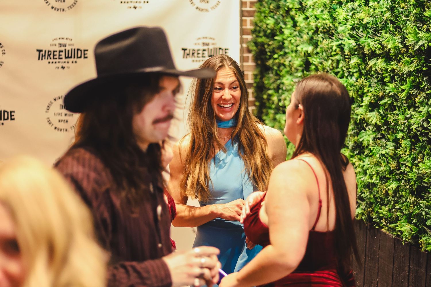 People socializing and laughing at a Three Wide Brewing Co. event in front of a green ivy wall backdrop.