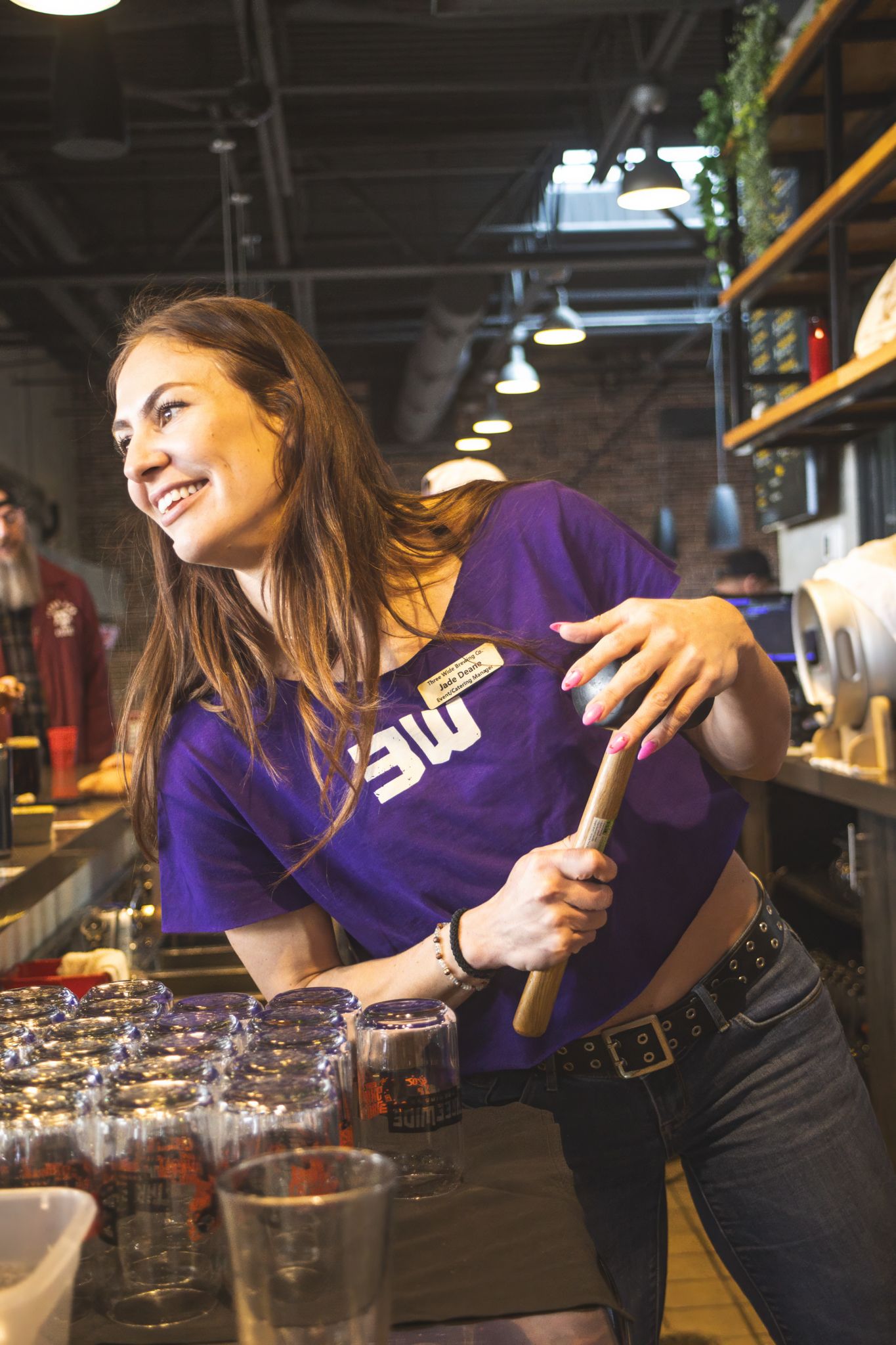 Cheerful woman in purple UW shirt holds a wooden mallet near glasses at a bar counter.