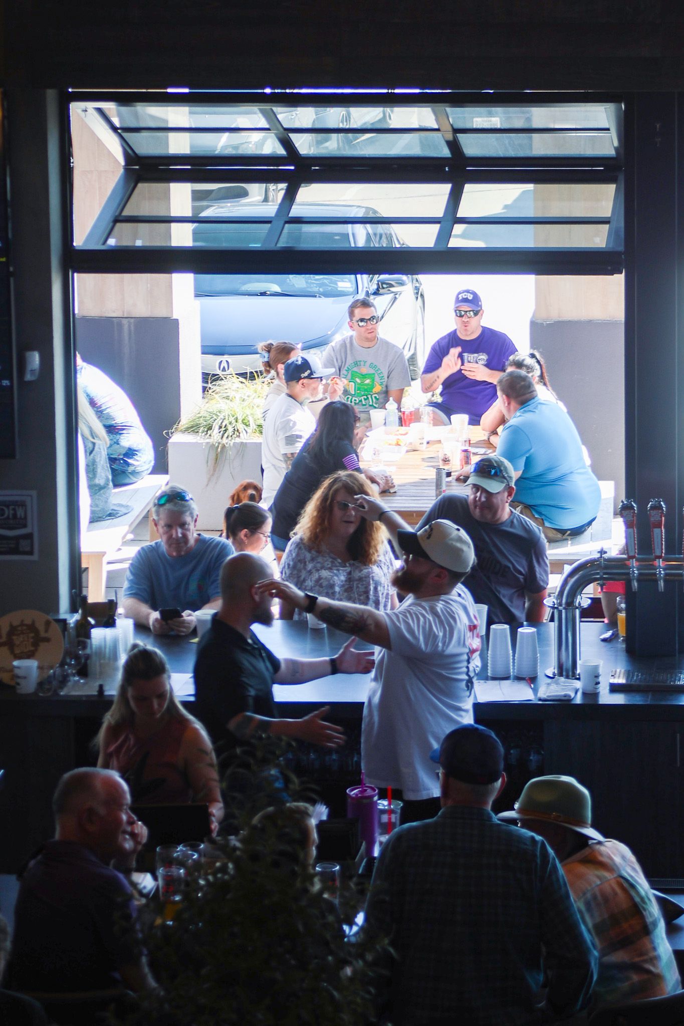 Busy bar scene with patrons socializing indoors and outdoors through large open garage-style window.