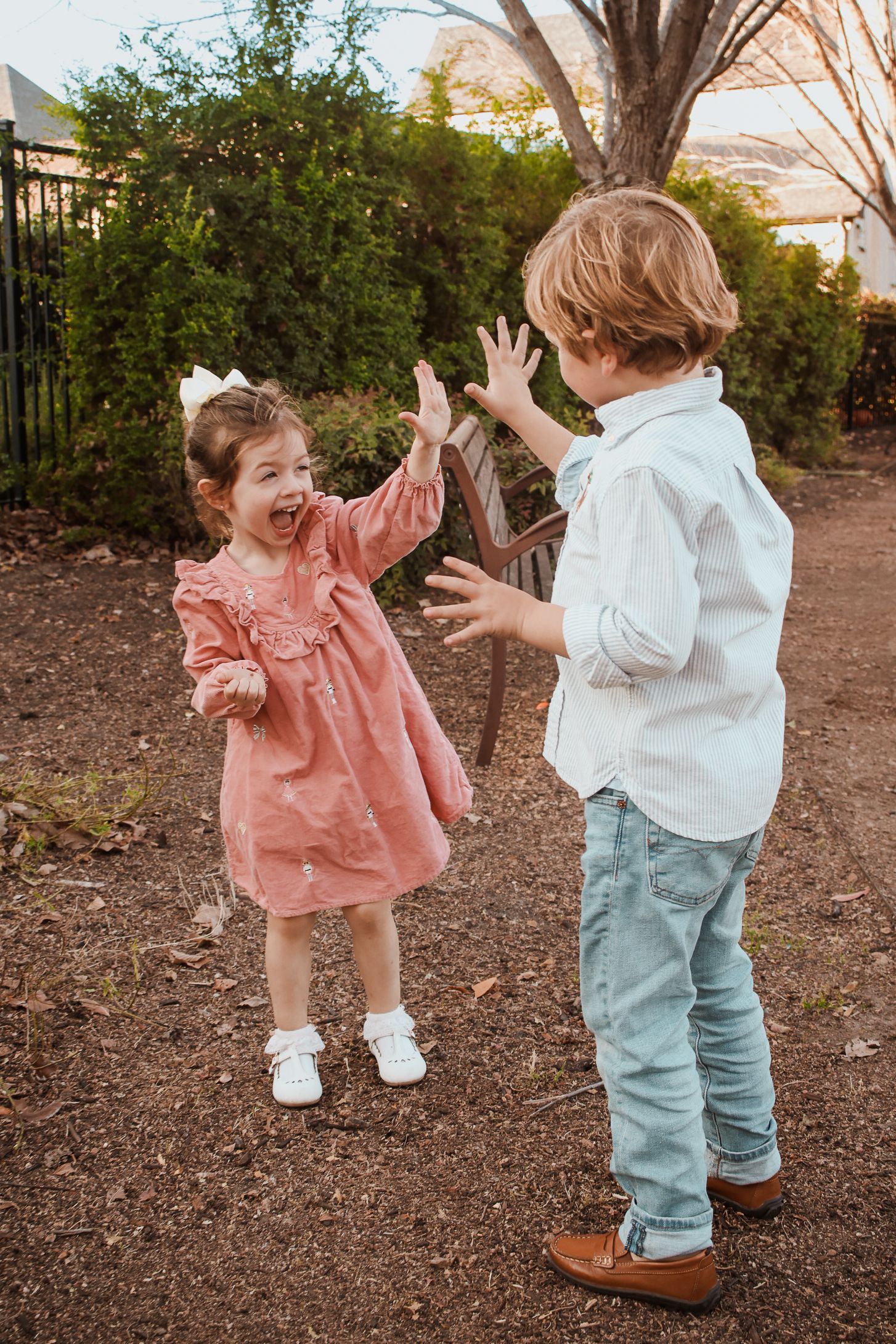 Two young children playing outdoors, a girl in a pink dress and a boy in jeans giving a high five.