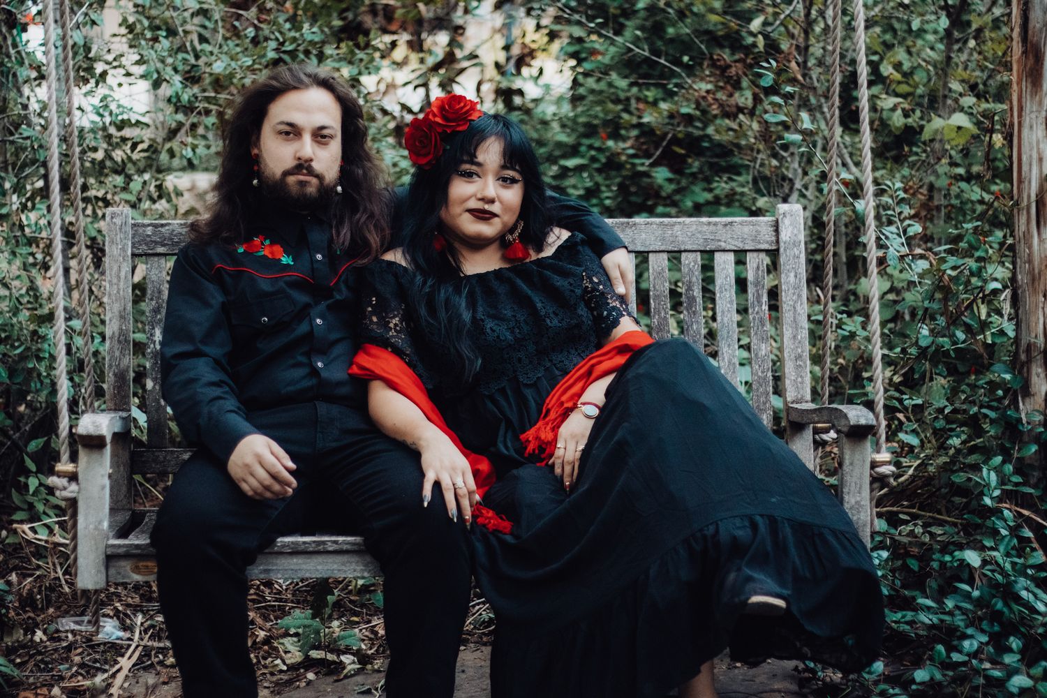 Couple in matching black and red gothic outfits sitting on a wooden bench surrounded by lush greenery.