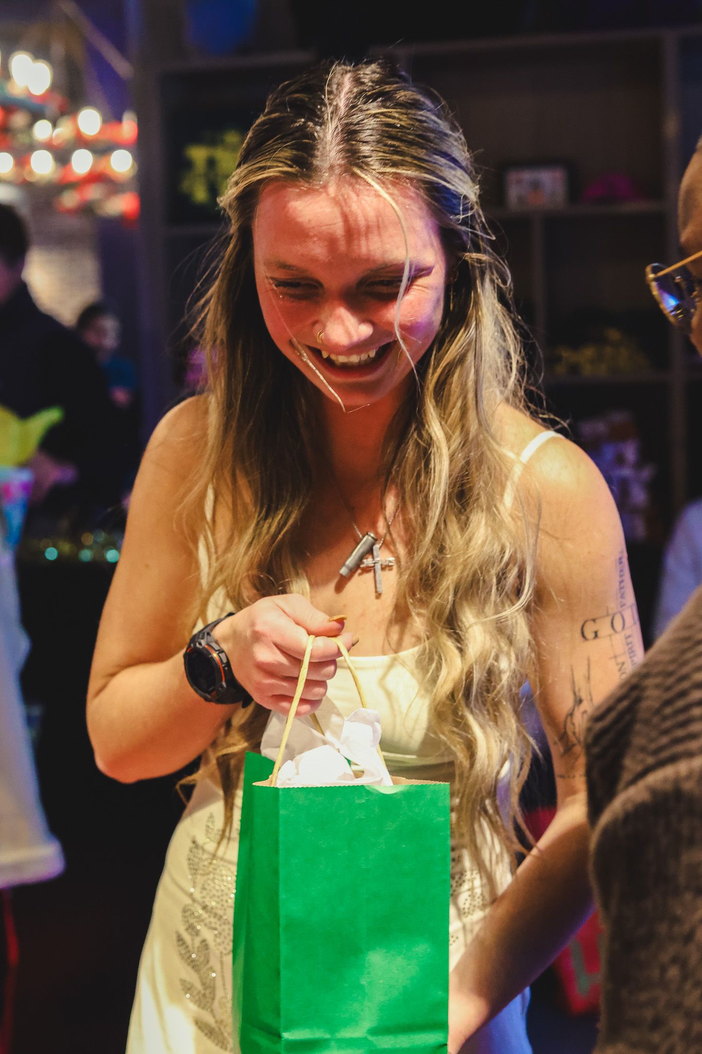 Smiling woman with long blonde hair opening a green gift bag at a party.
