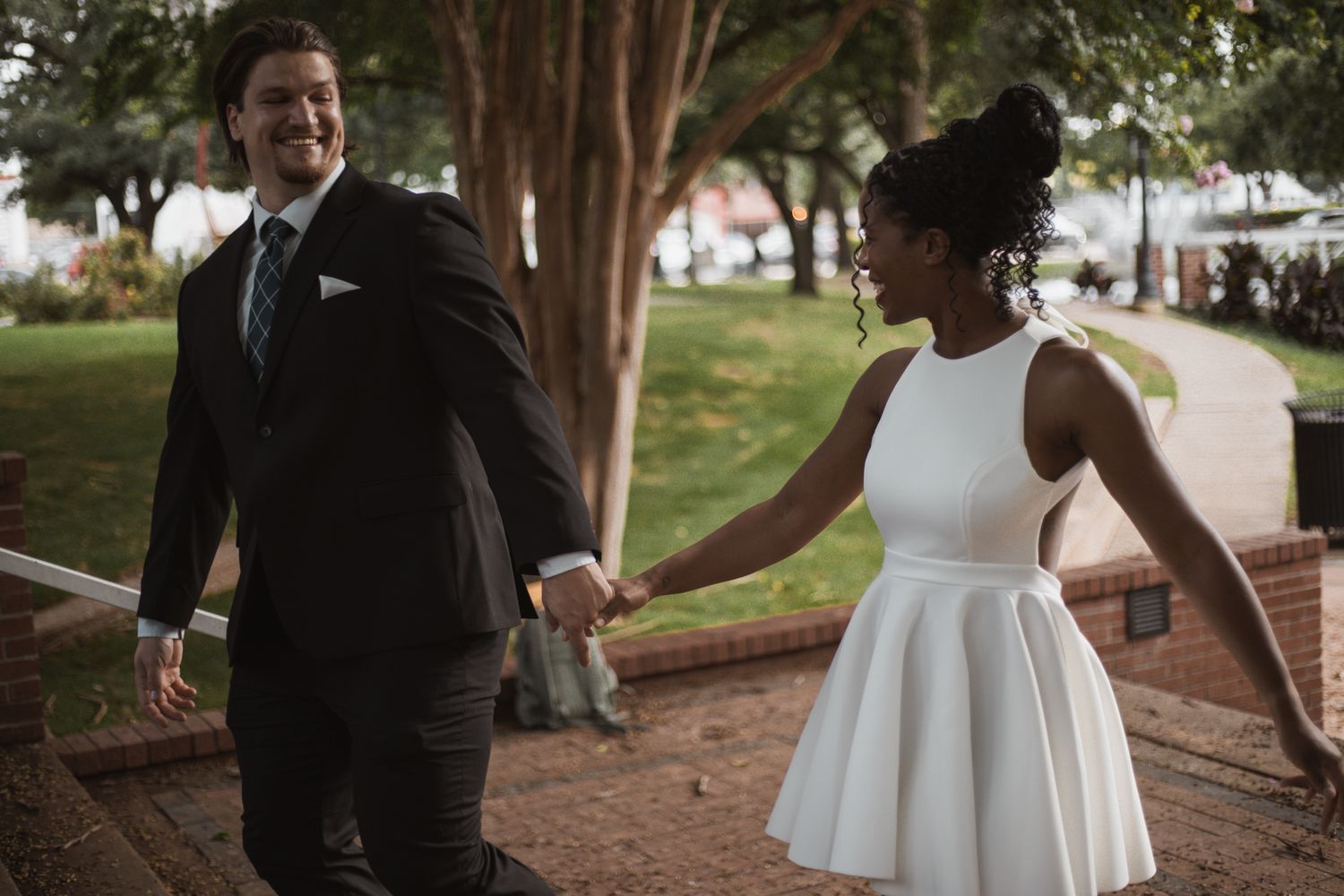 Interracial couple holding hands in wedding attire, bride in white mini dress, groom in dark suit, outdoor brick path.