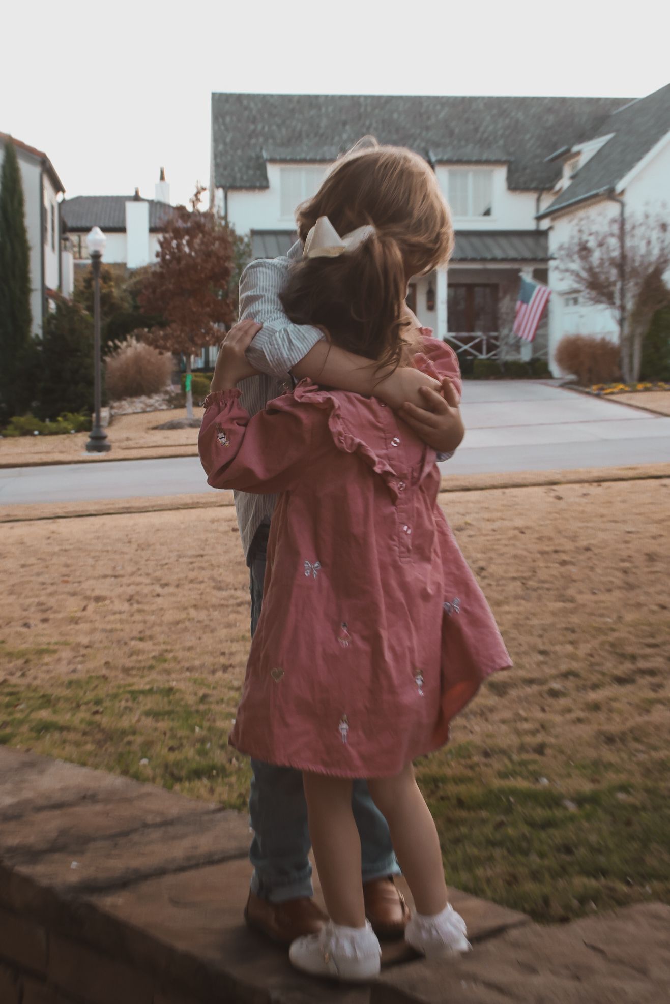 Two young girls hugging outdoors in a suburban neighborhood, one wearing a pink dress with a bow in her hair.