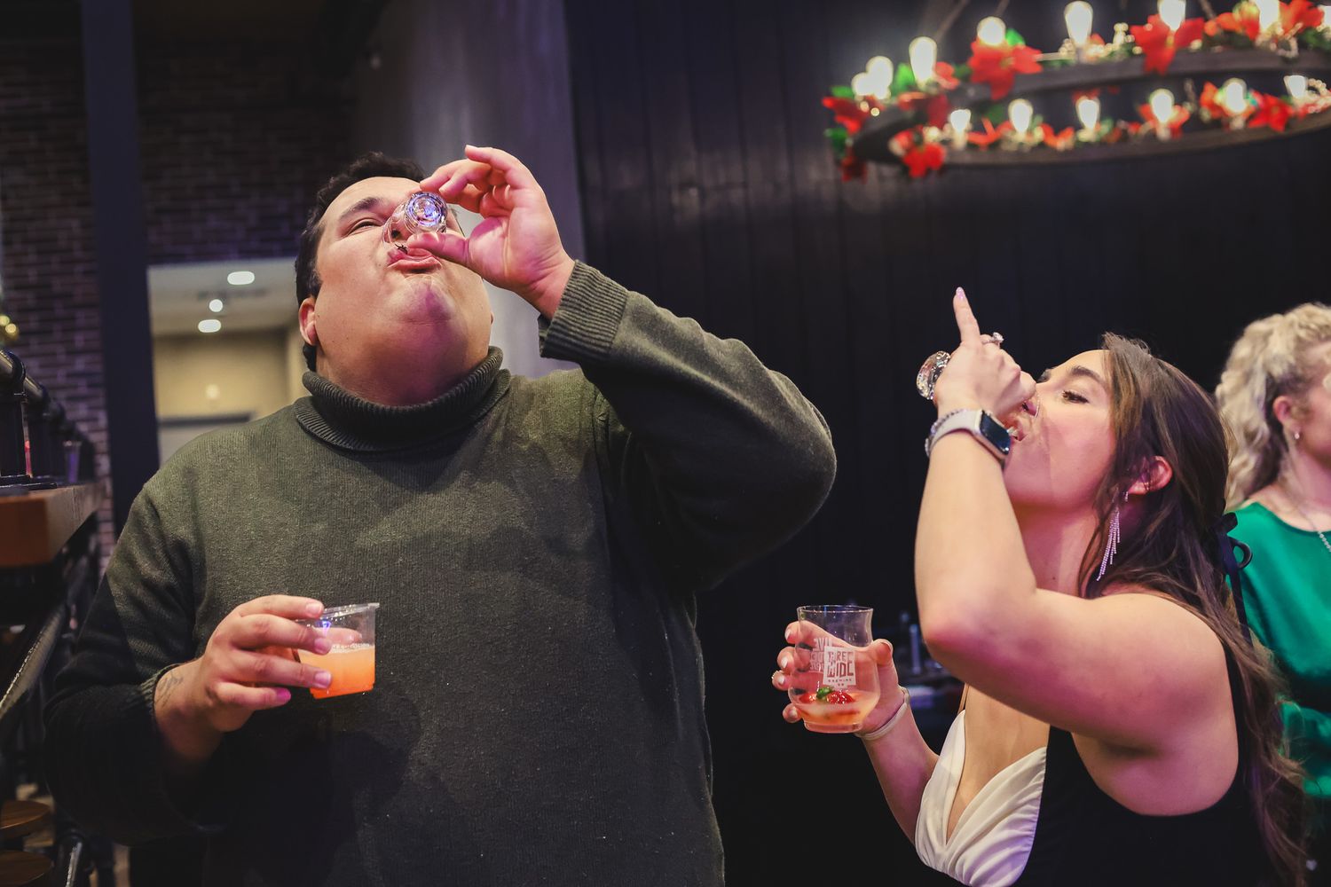 Man and woman doing shots at a holiday party, each holding additional drinks in festive venue.