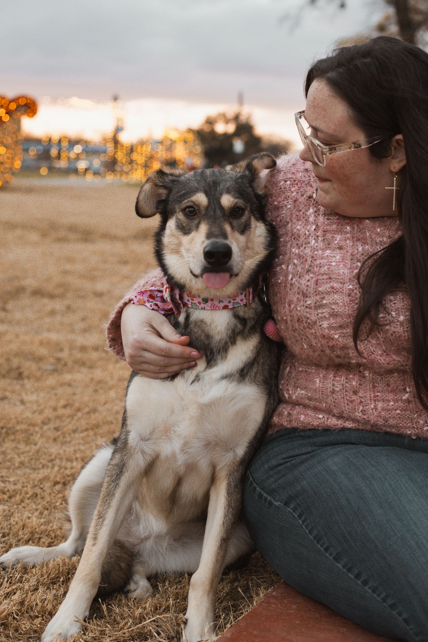 Woman in pink sweater sitting outdoors at dusk with her husky mix dog wearing a pink harness.