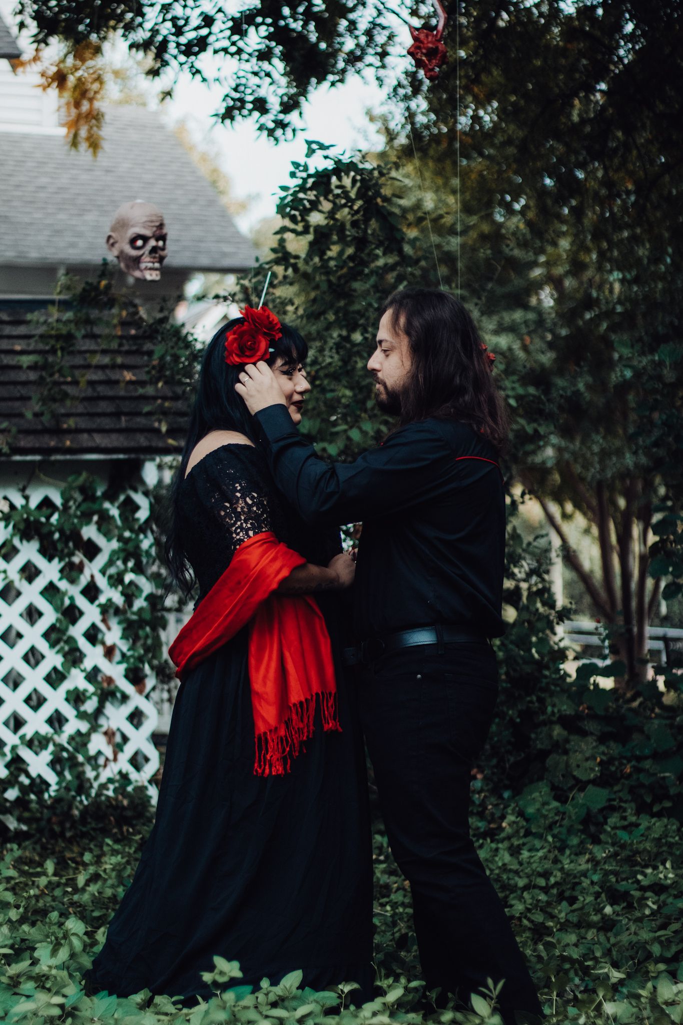 Couple in black Gothic Halloween costumes dancing outdoors with red scarf and skull decoration among green trees.