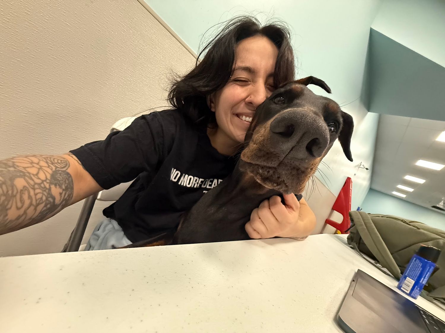 Smiling woman with tattoos taking a selfie with a large chocolate brown dog resting its head on a desk.