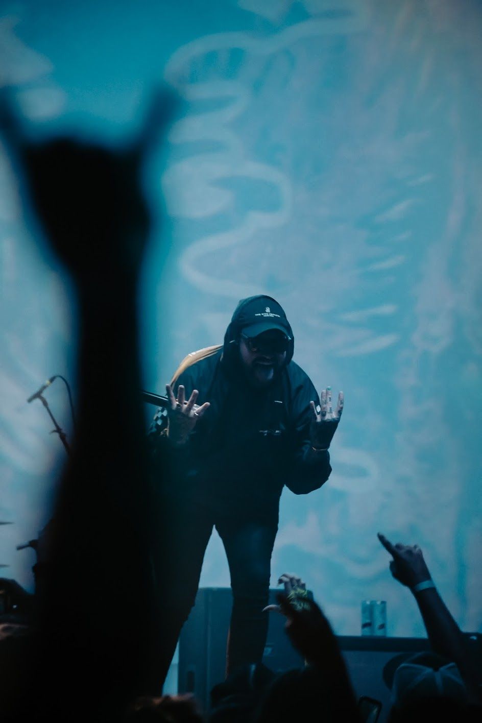 Concert performer on stage gesturing to crowd with blue atmospheric lighting and raised hands.