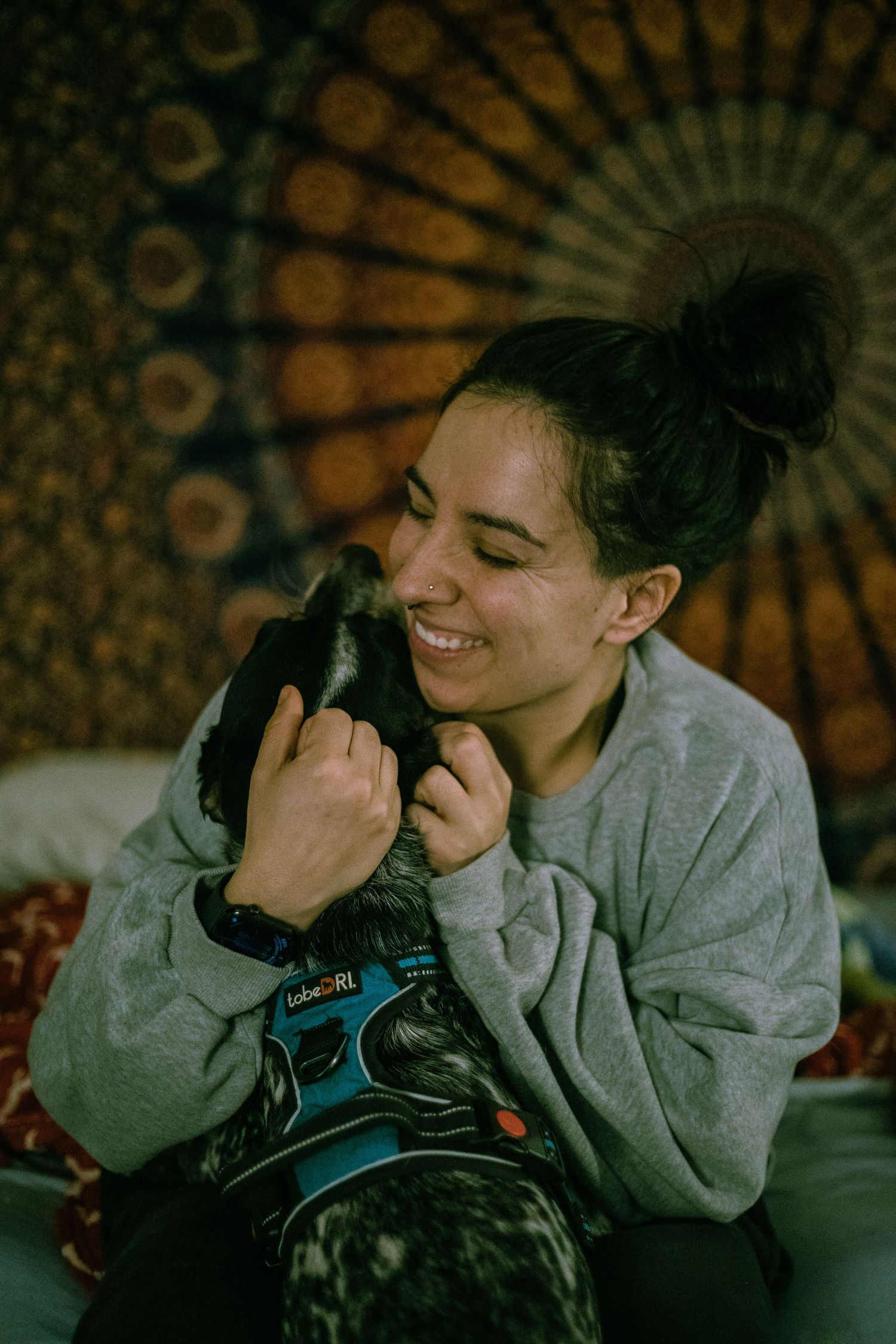 Smiling woman in grey hoodie cuddling her black and white dog against a colorful tapestry backdrop.