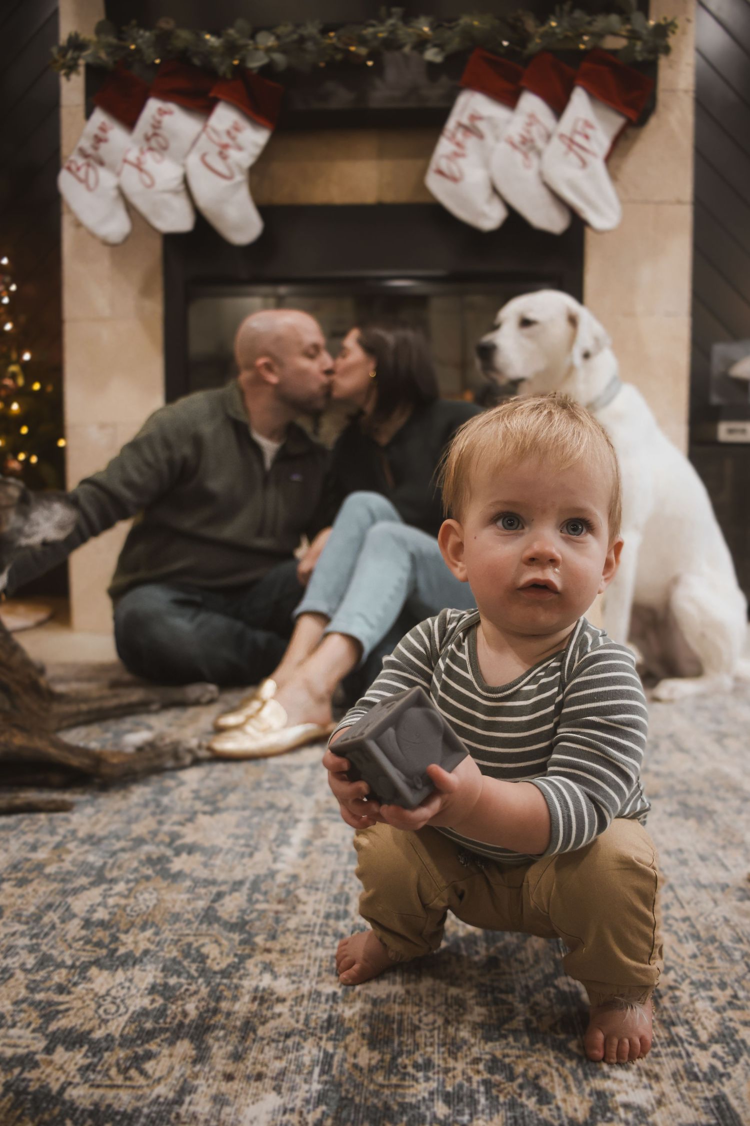 Toddler plays with toy in front of parents kissing by Christmas fireplace with white dog nearby.