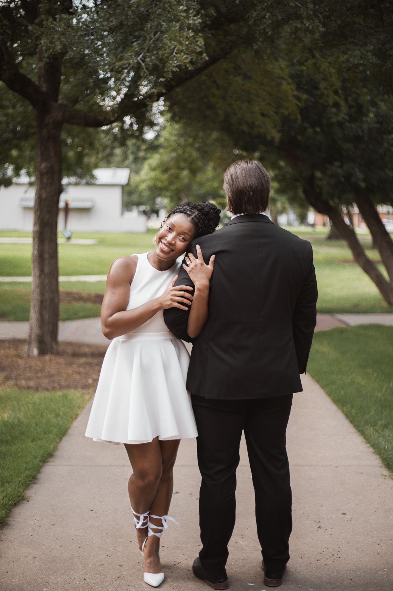A smiling bride in a white dress embraces her groom in a black suit on a tree-lined sidewalk.
