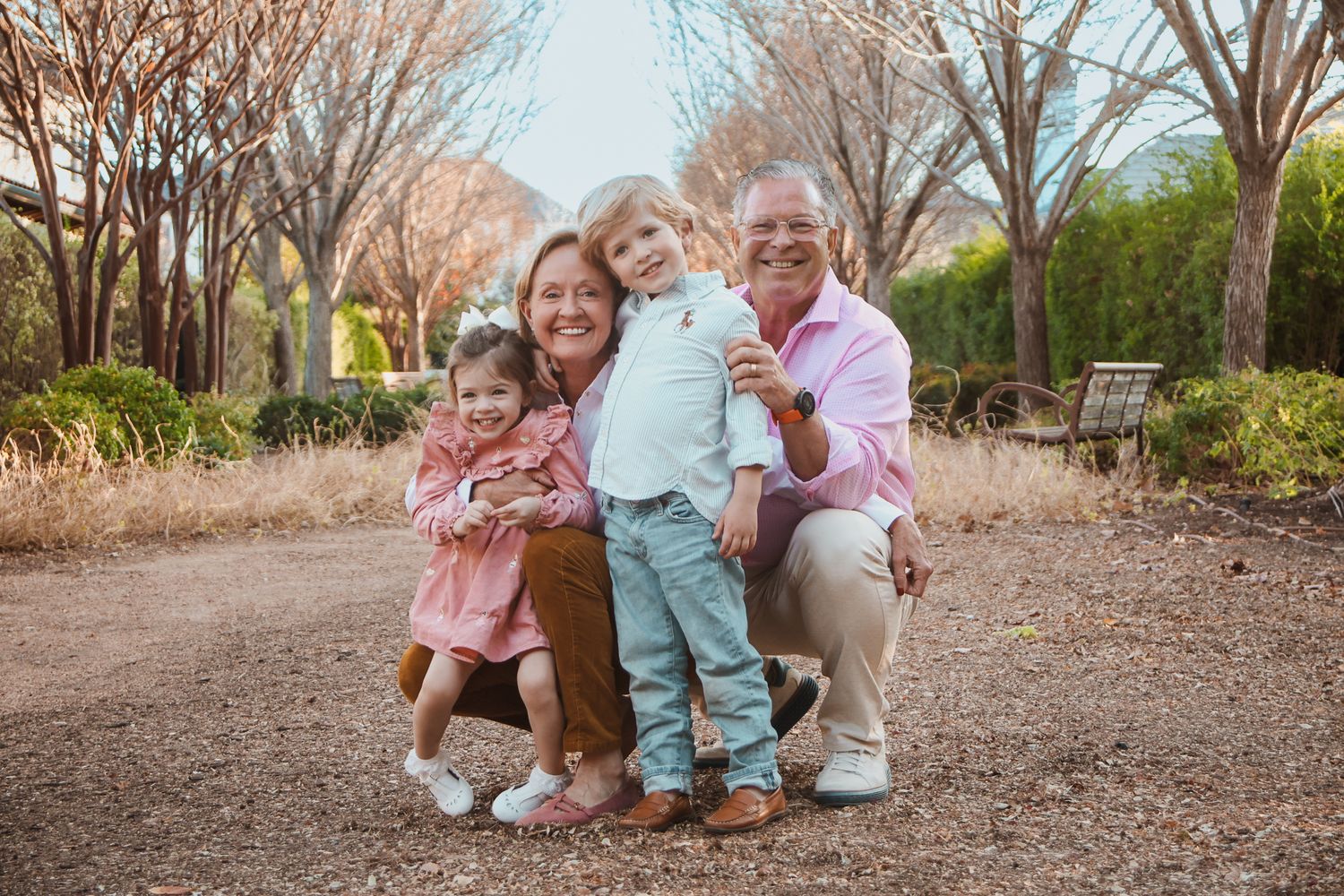 Family of four posing outdoors in a park, grandparents with two young children smiling in pink and pastel outfits.