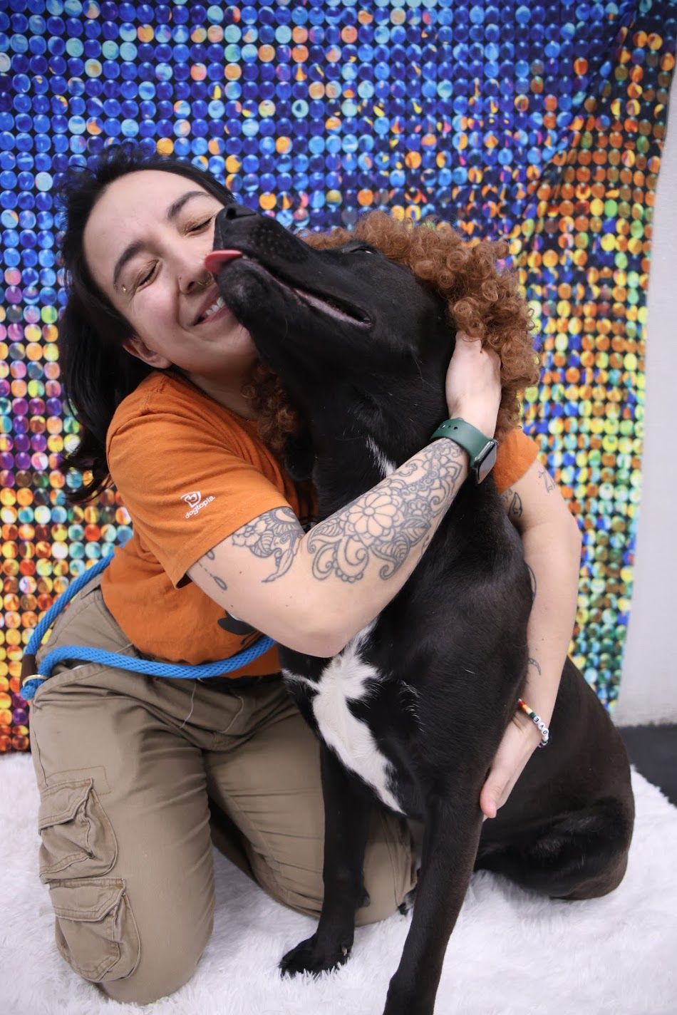 Tattooed woman in orange shirt being licked by a black and white dog on white rug.