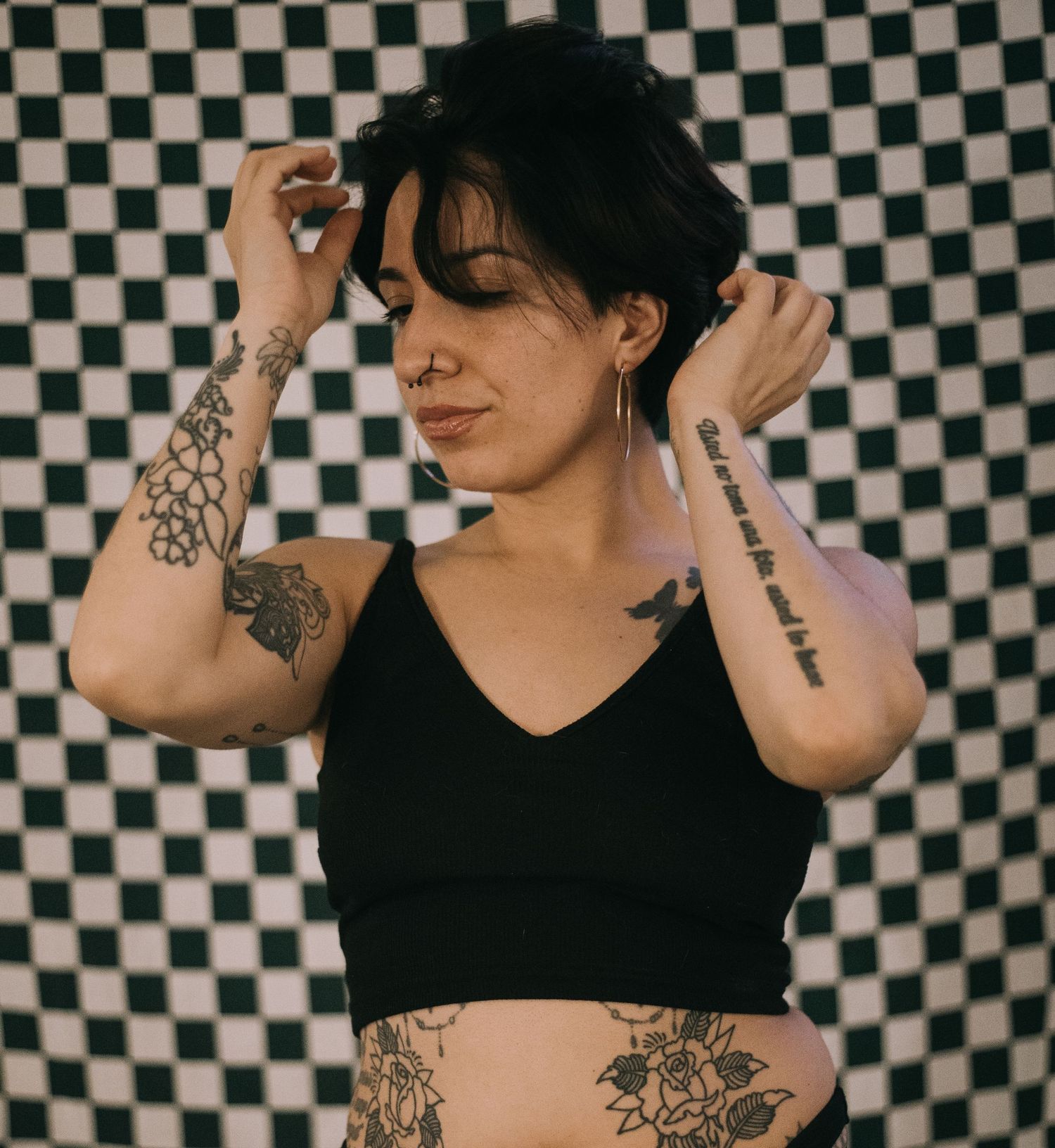 Tattooed woman in black crop top posing against checkered background, with visible arm and stomach tattoos.