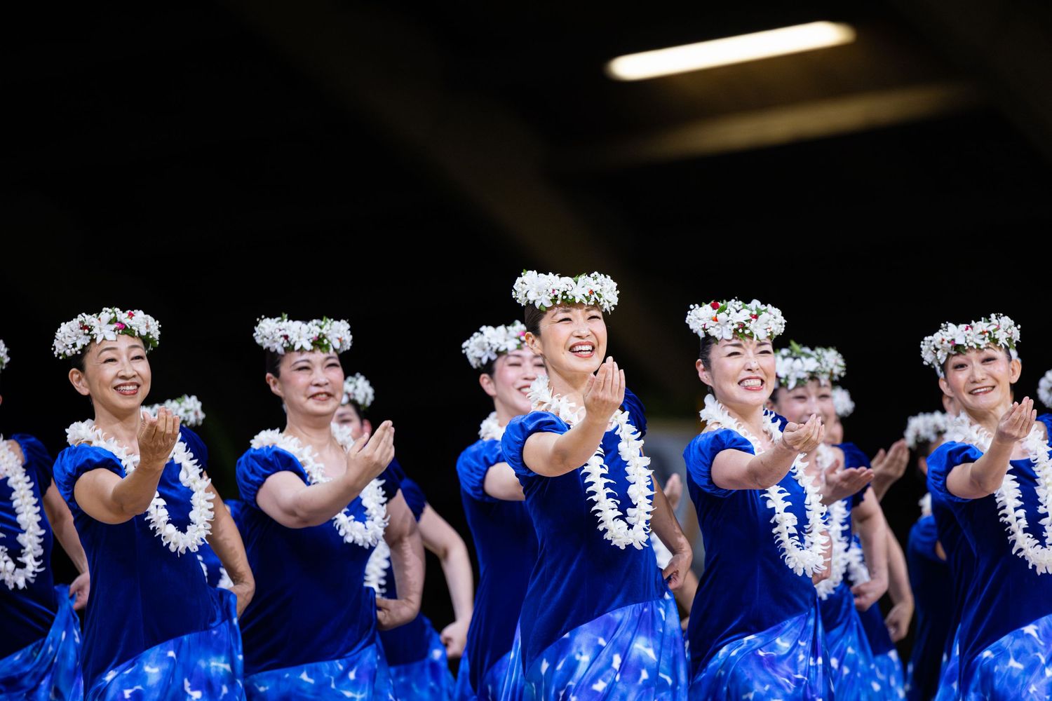 A group of performers wearing bright blue outfits and white leis dance together in synchronized formation.