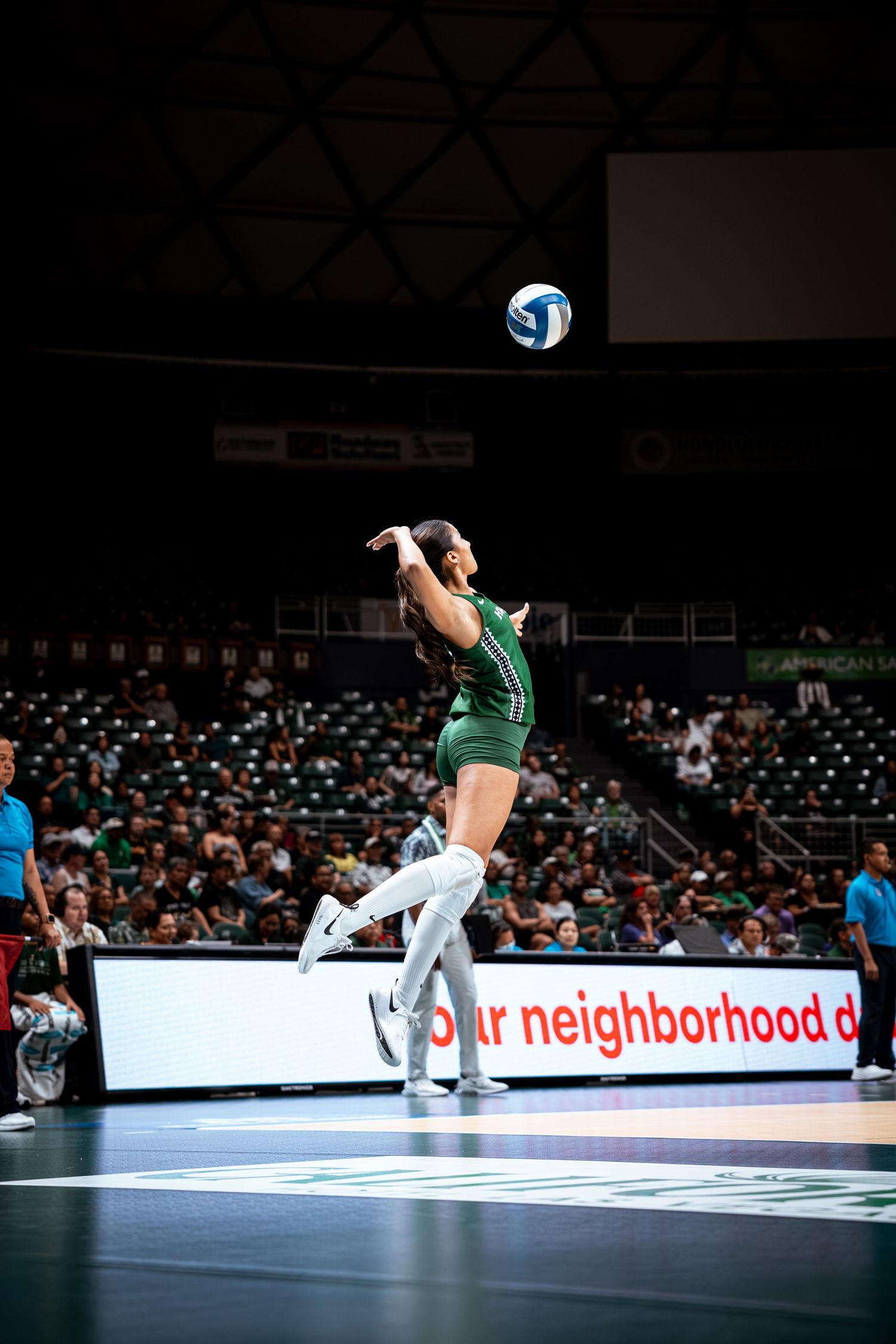 Female volleyball player in green uniform jumps to serve during an indoor arena match.