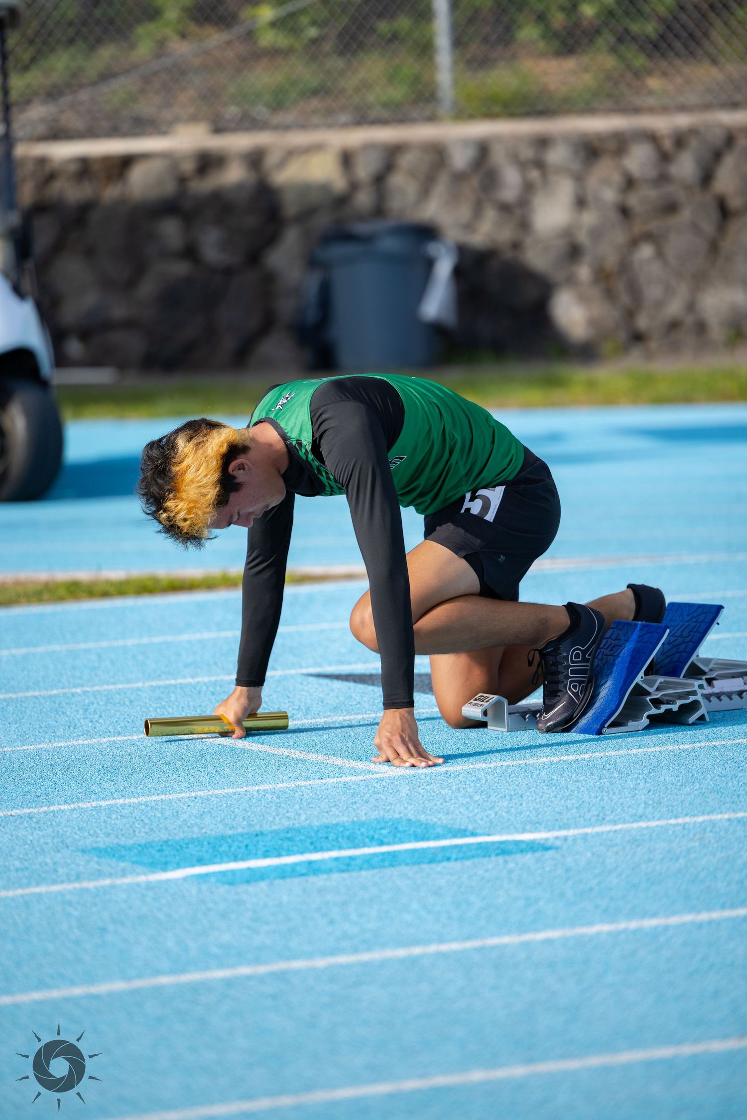 Runner in green uniform crouching at starting position on blue track.