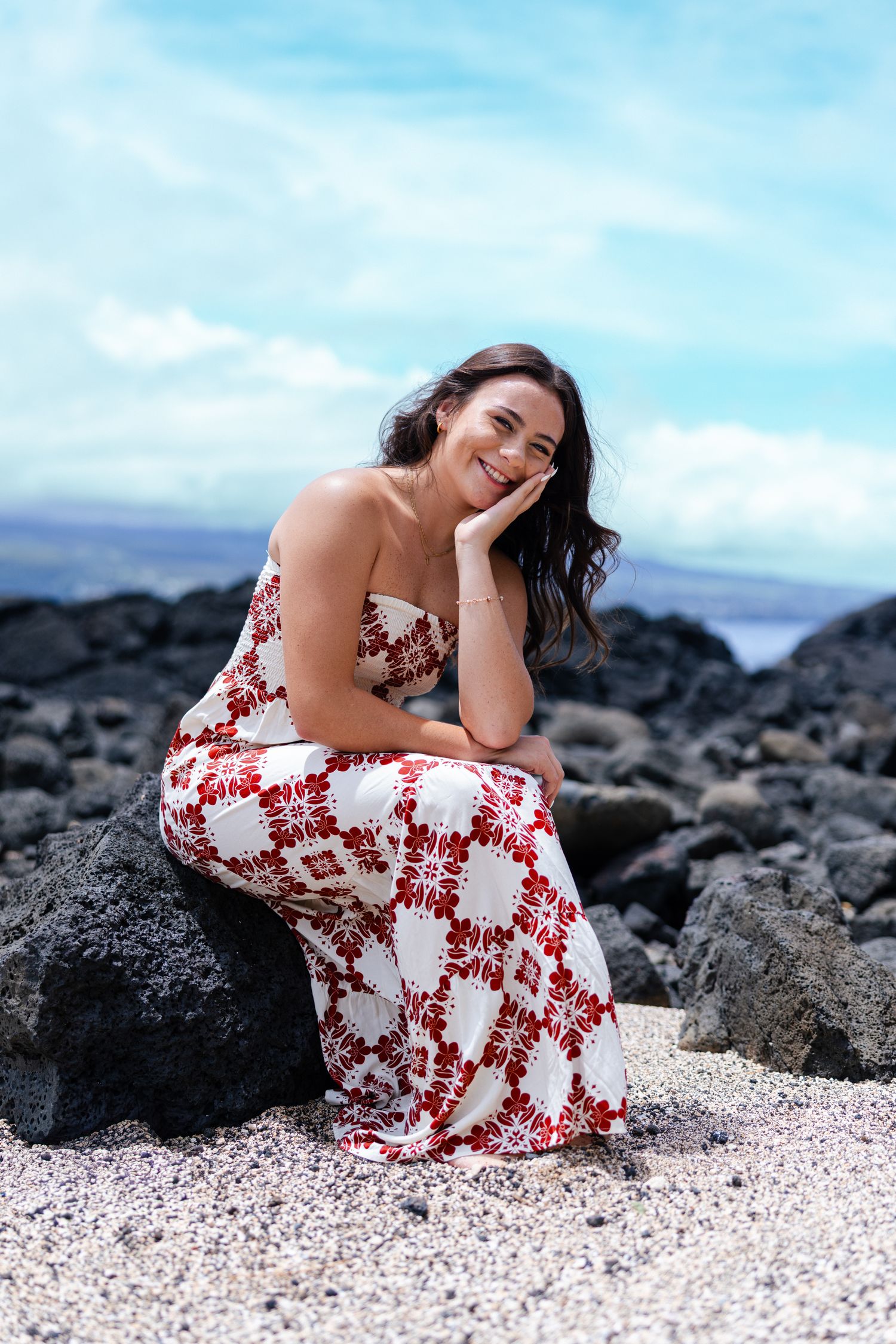 Smiling woman in red floral maxi dress sitting on volcanic rocks at a Hawaiian beach with blue sky backdrop.