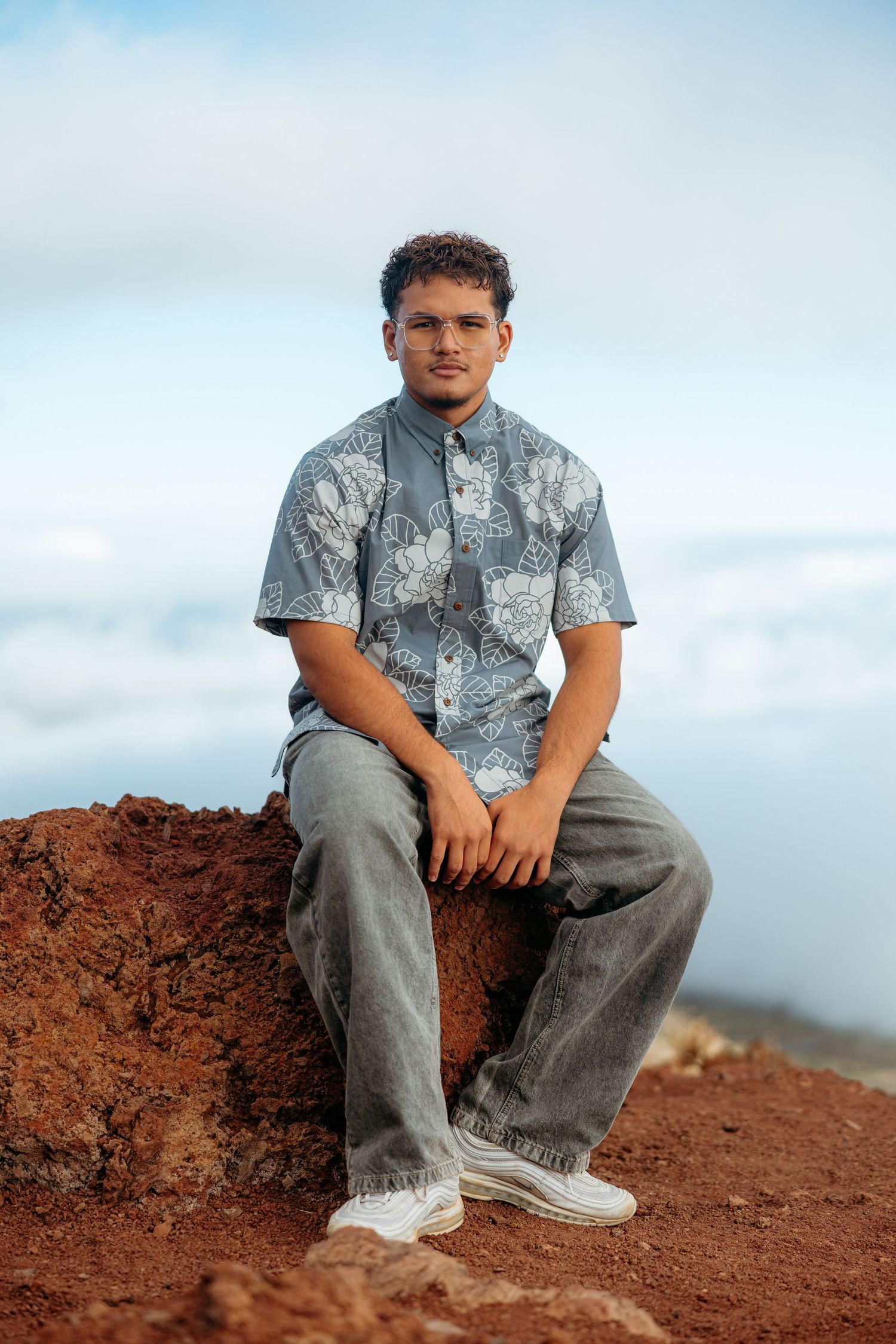 A person in a gray patterned shirt and jeans sits on red volcanic soil against a cloudy sky.