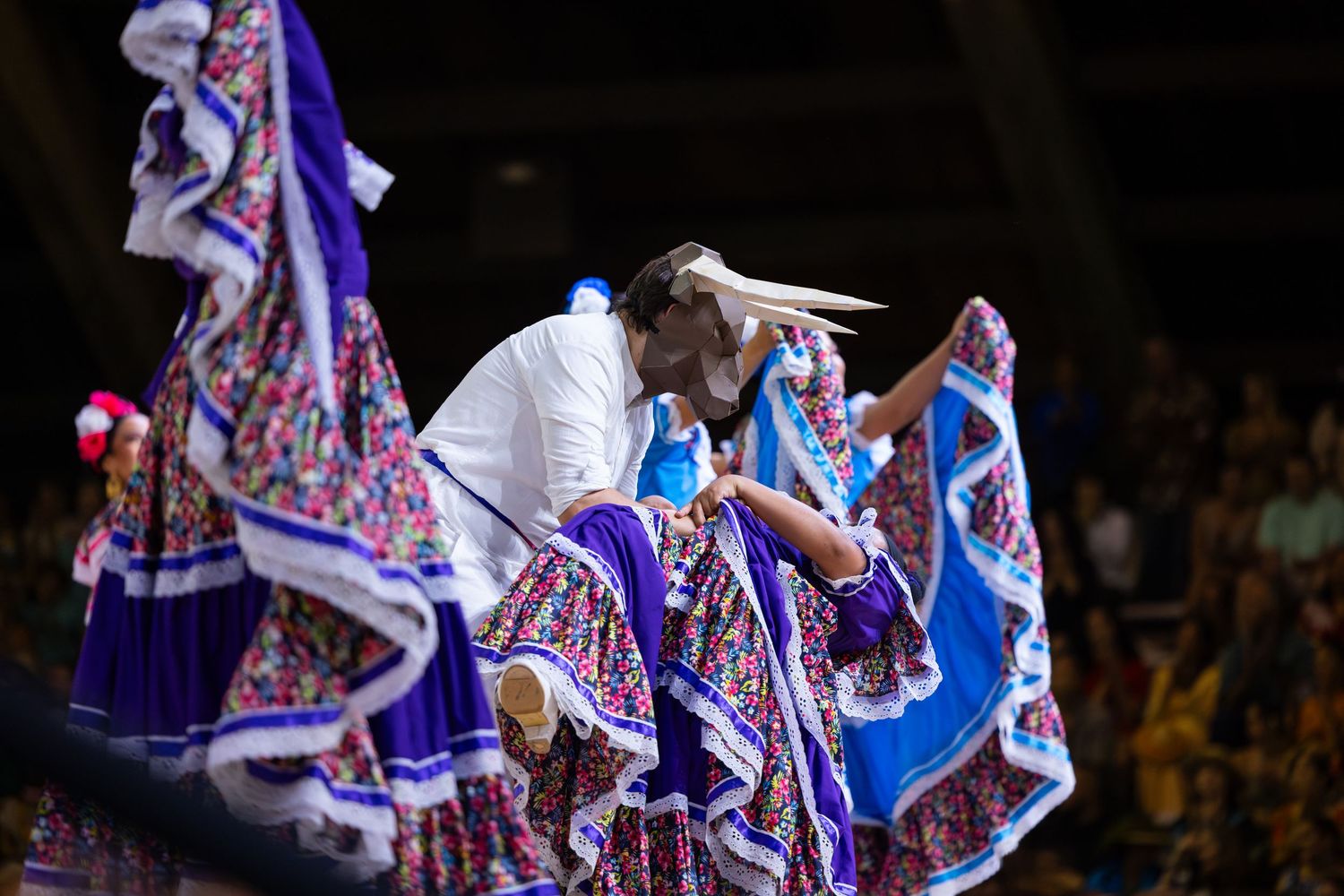 Dancers in flowing blue and white traditional dresses perform a vibrant folk dance on stage.