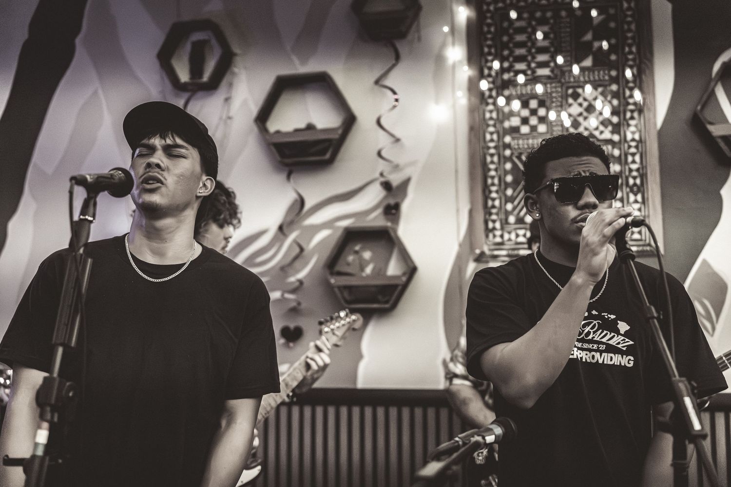 Two performers on stage in black and white photo with one singing into microphone and one wearing sunglasses.