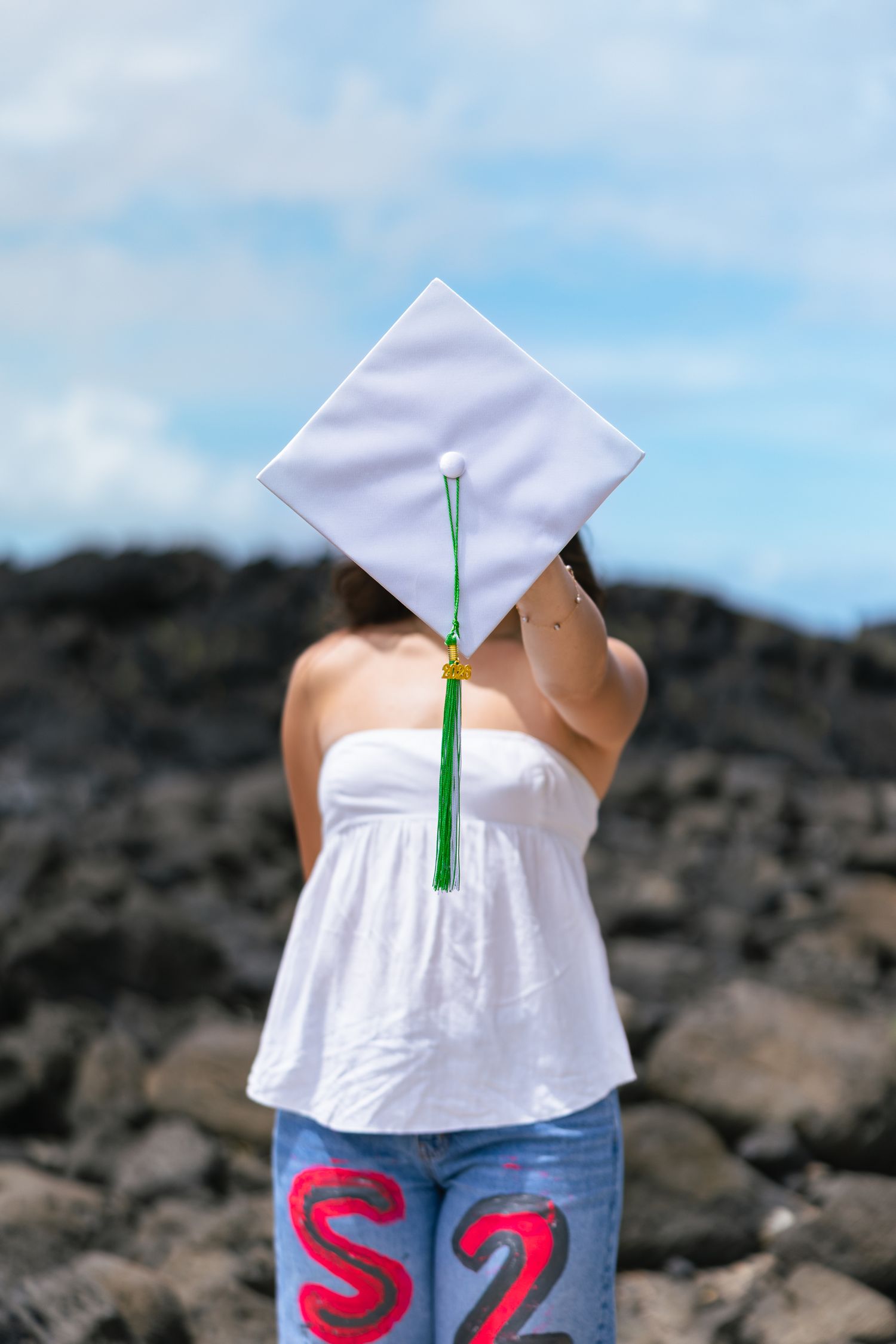 A graduate holds a white mortarboard cap with green tassel in front of her face outdoors near rocky terrain.