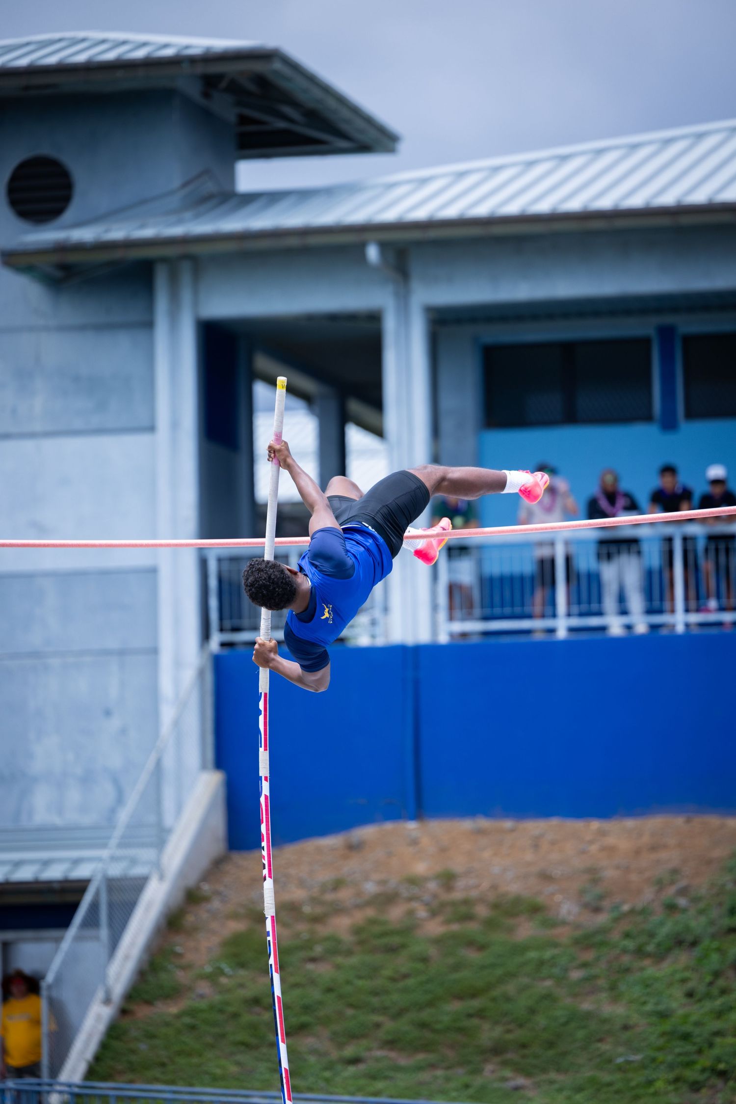 Athlete performing pole vault jump against blue sky and buildings.