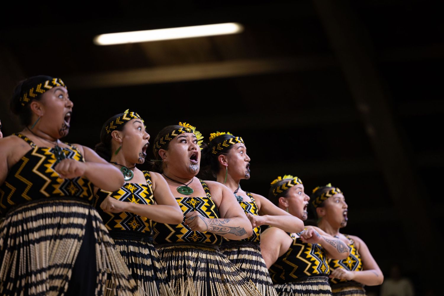 Traditional Maori performers wearing traditional dress and floral headpieces perform in unison on stage.