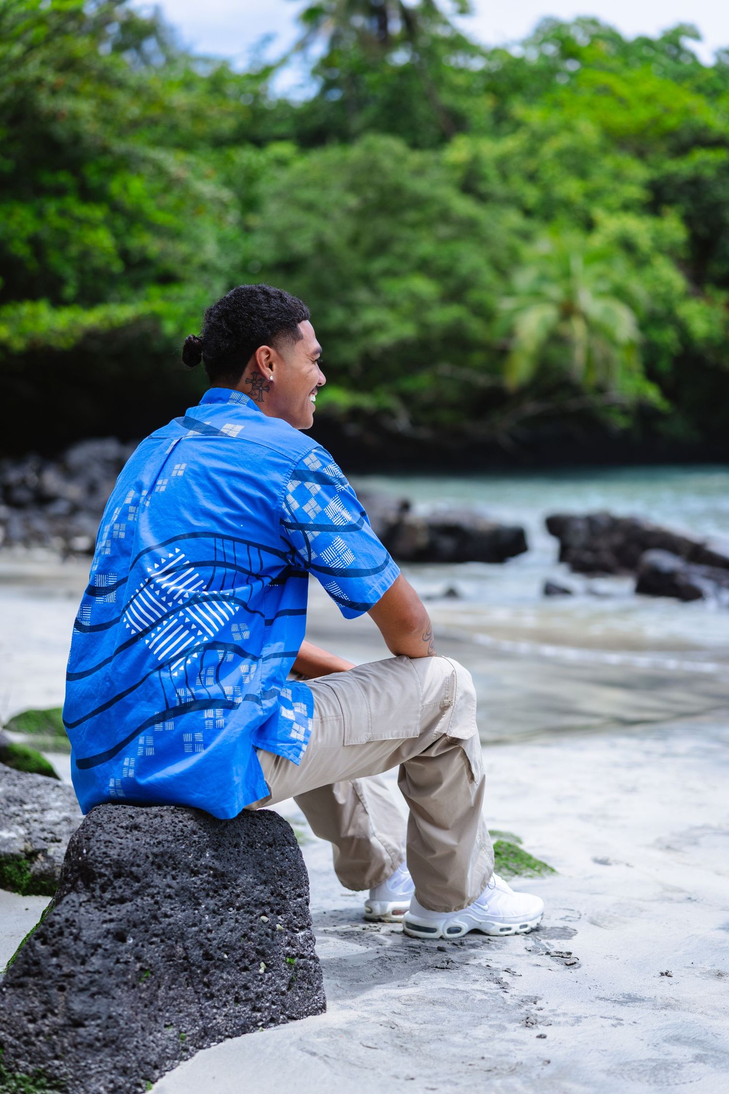 Young man in blue Hawaiian shirt sitting on rock at tropical beach, smiling and looking away.