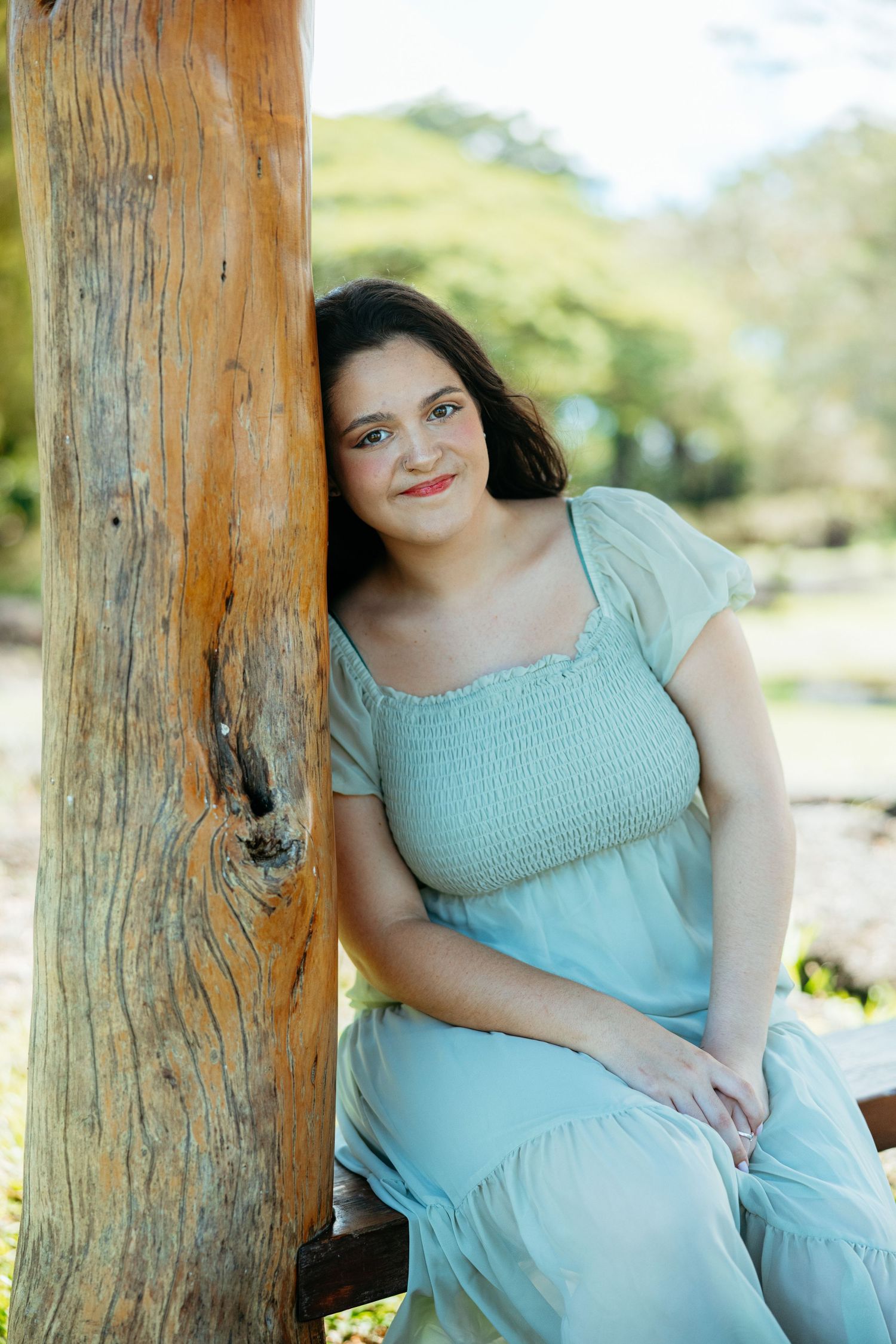 A person in a light turquoise dress poses against a wooden post outdoors in a natural setting.