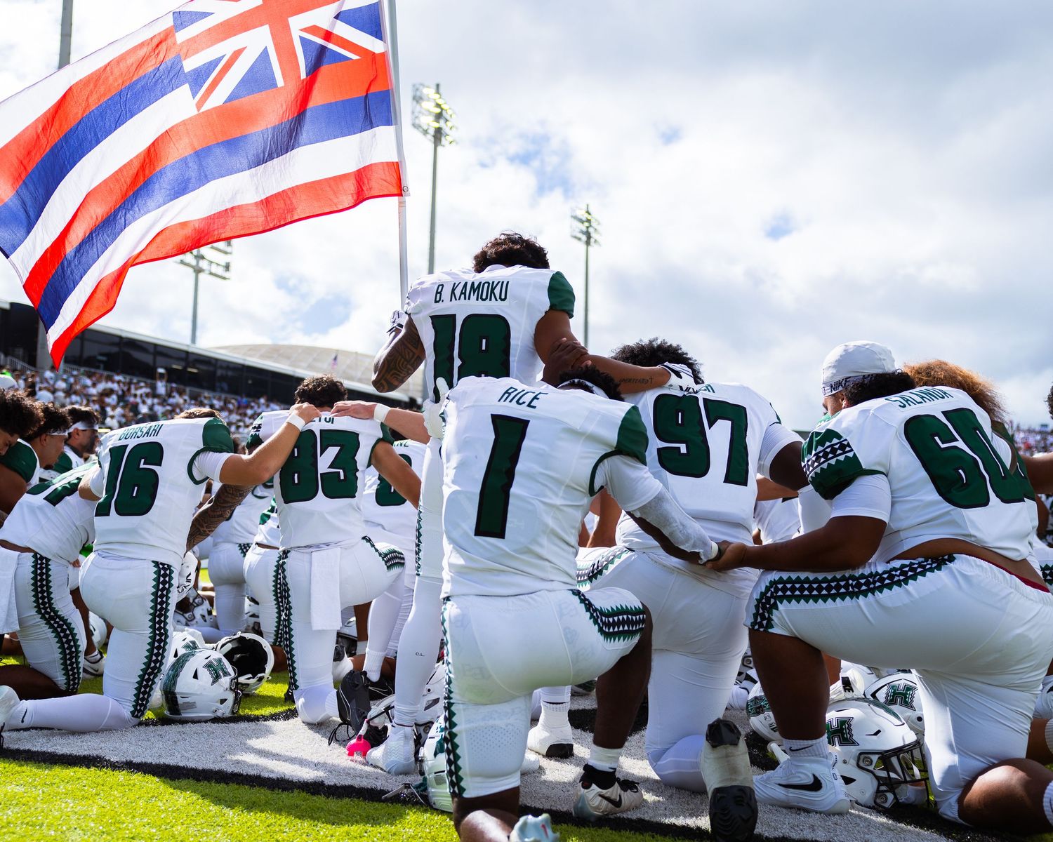 University of Hawaii Warriors football players kneel together on the sideline with the Hawaii state flag waving above.