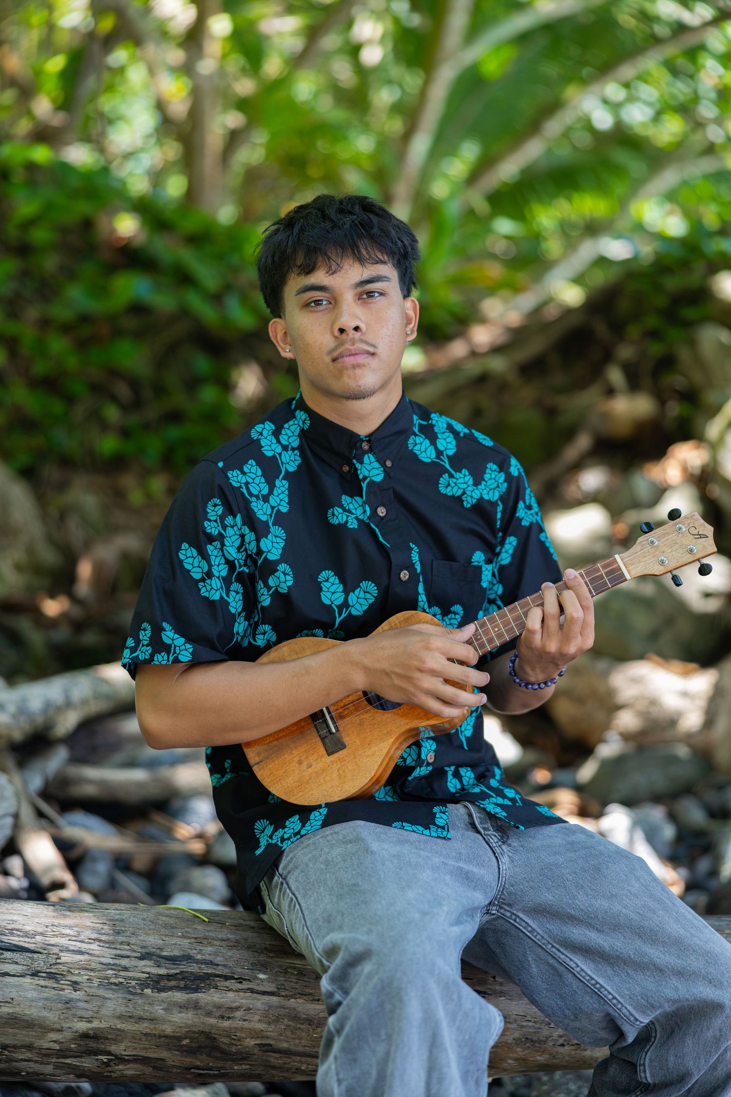 A musician in a black and turquoise floral shirt plays ukulele while sitting on a fallen log in a forest setting.