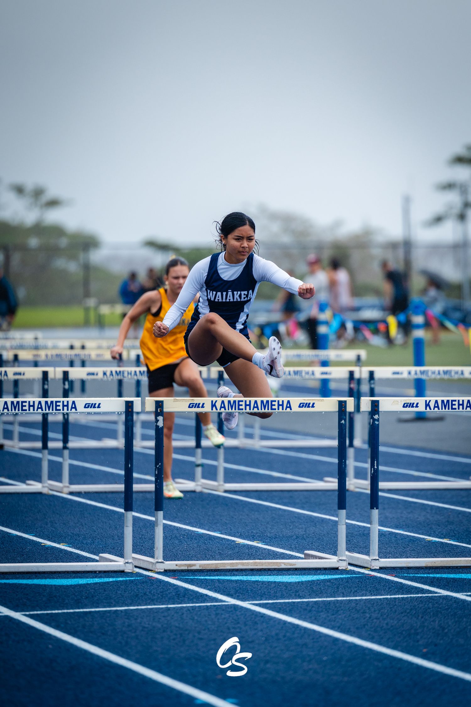High school track athlete clears hurdles during competition on blue running track at Kamehameha meet.