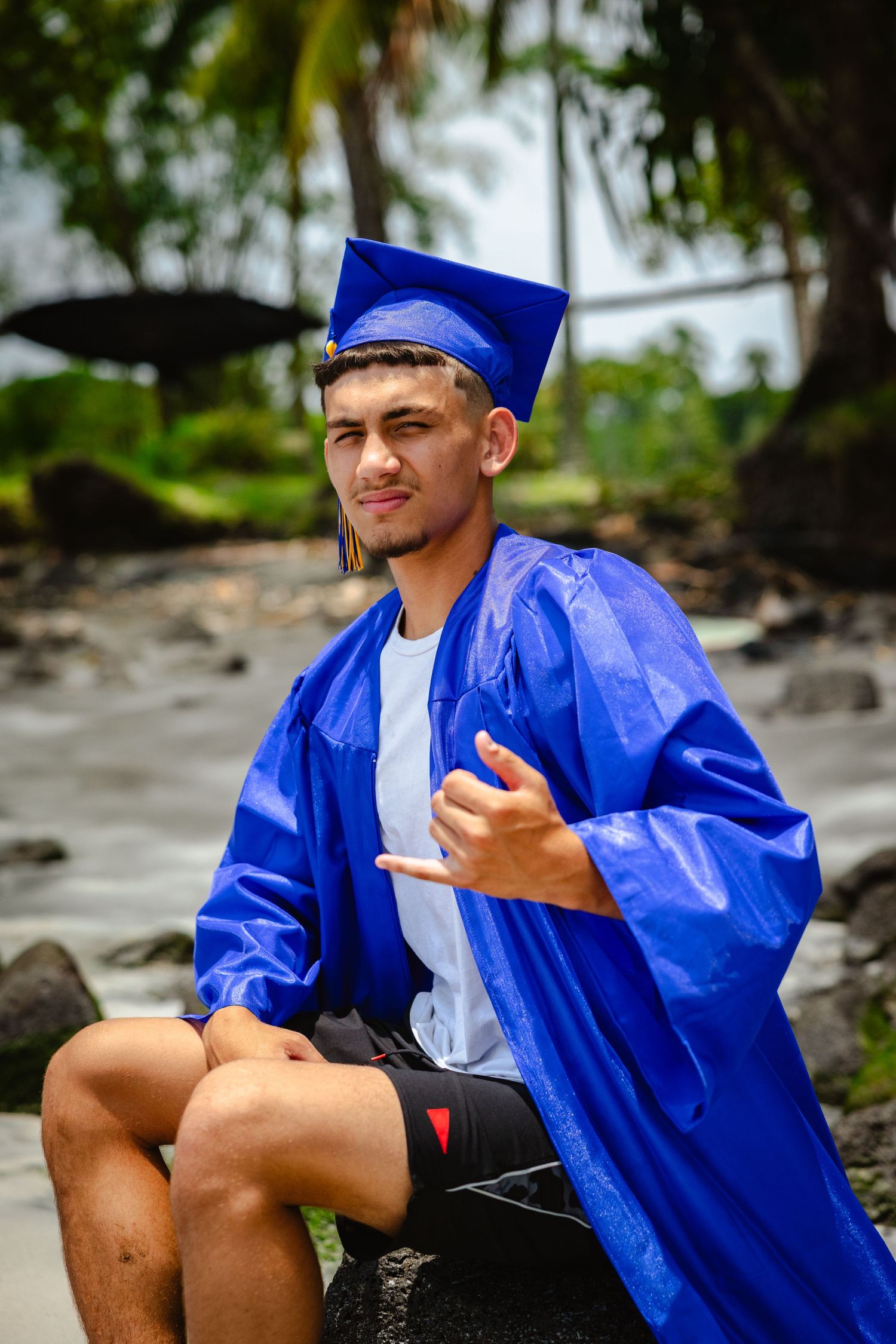 Young man in blue graduation cap and gown sitting outdoors on rocks with tropical trees in background.