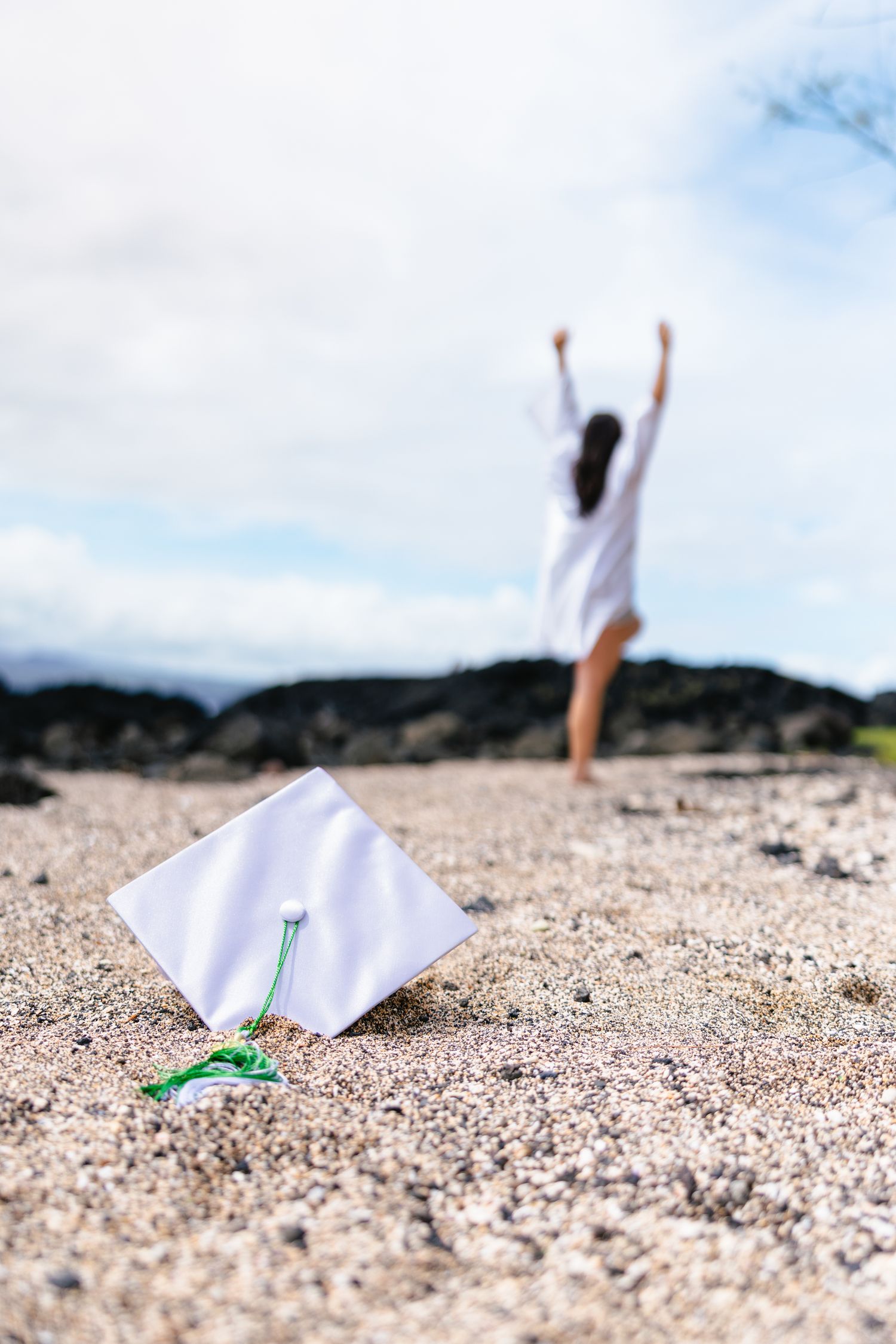 White graduation cap resting on sandy beach with celebrating graduate raising arms in background.