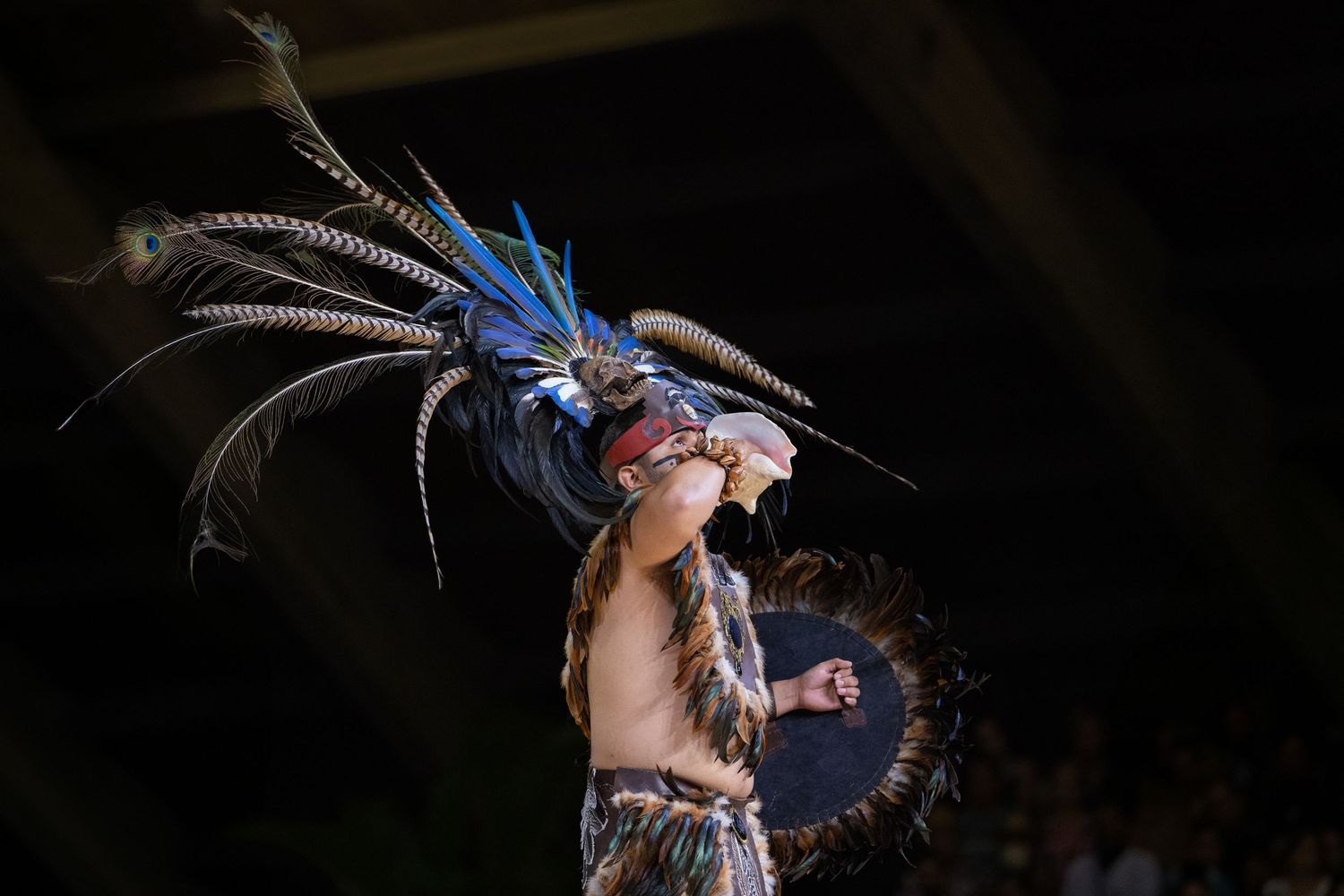 A performer in traditional Aztec ceremonial costume with blue feathered headdress dances during a cultural performance.