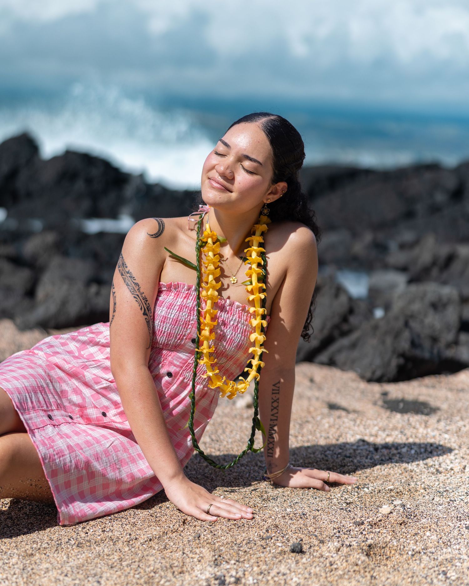 Person in pink dress lying on beach sand with yellow lei necklace against ocean backdrop.