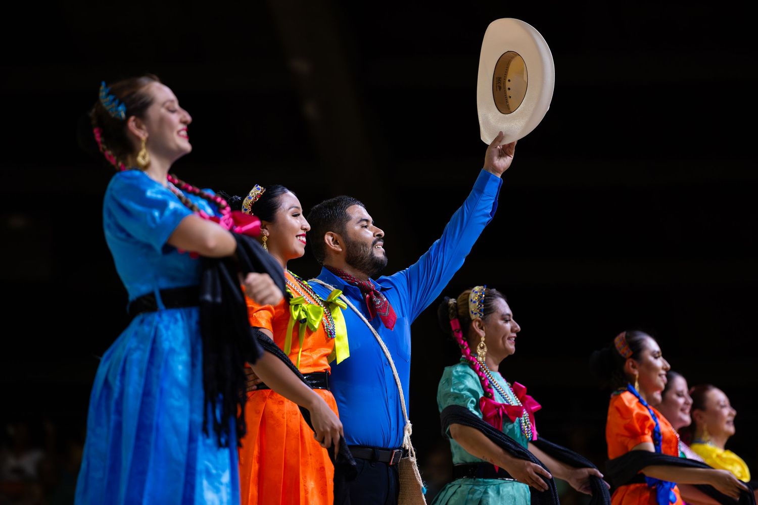 Performers in colorful outfits stand together on stage with arms raised during a theatrical performance.