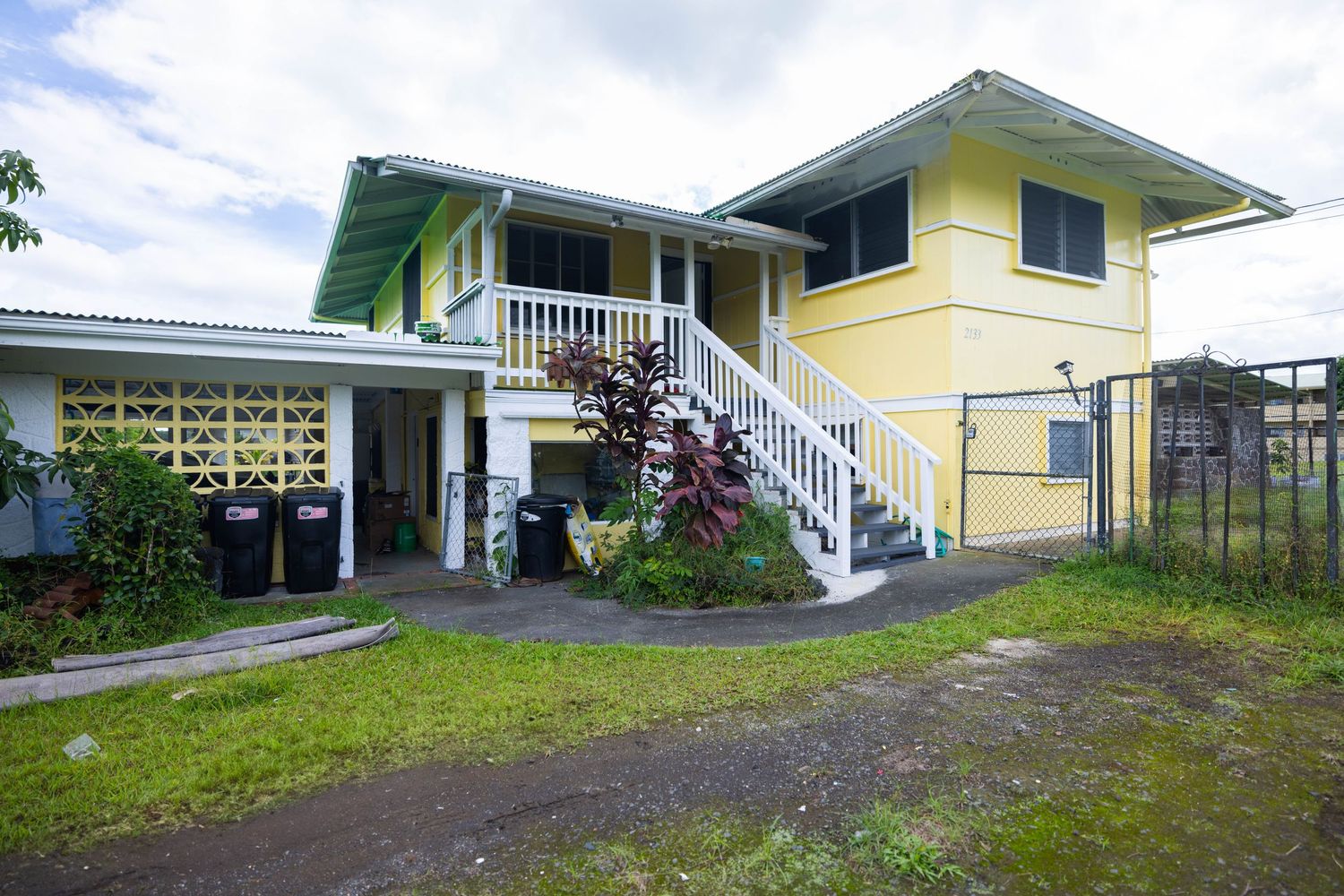 A panoramic view of a yellow Hawaiian-style house with white trim and a covered lanai surrounded by tropical landscaping.