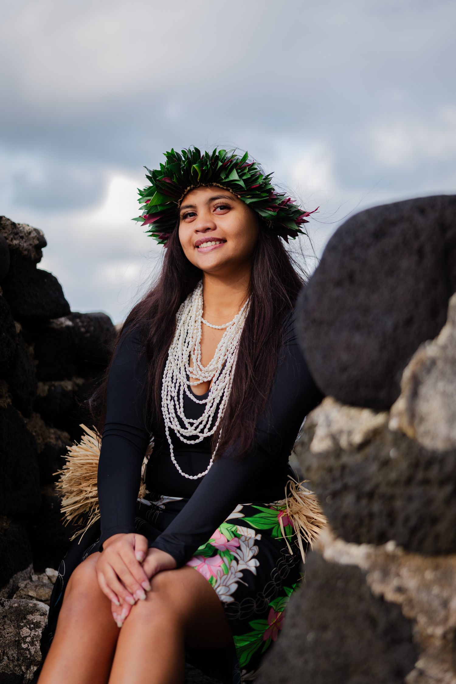 Young Hawaiian hula dancer in floral skirt, pearl lei, and leaf crown sits smiling among lava rocks.