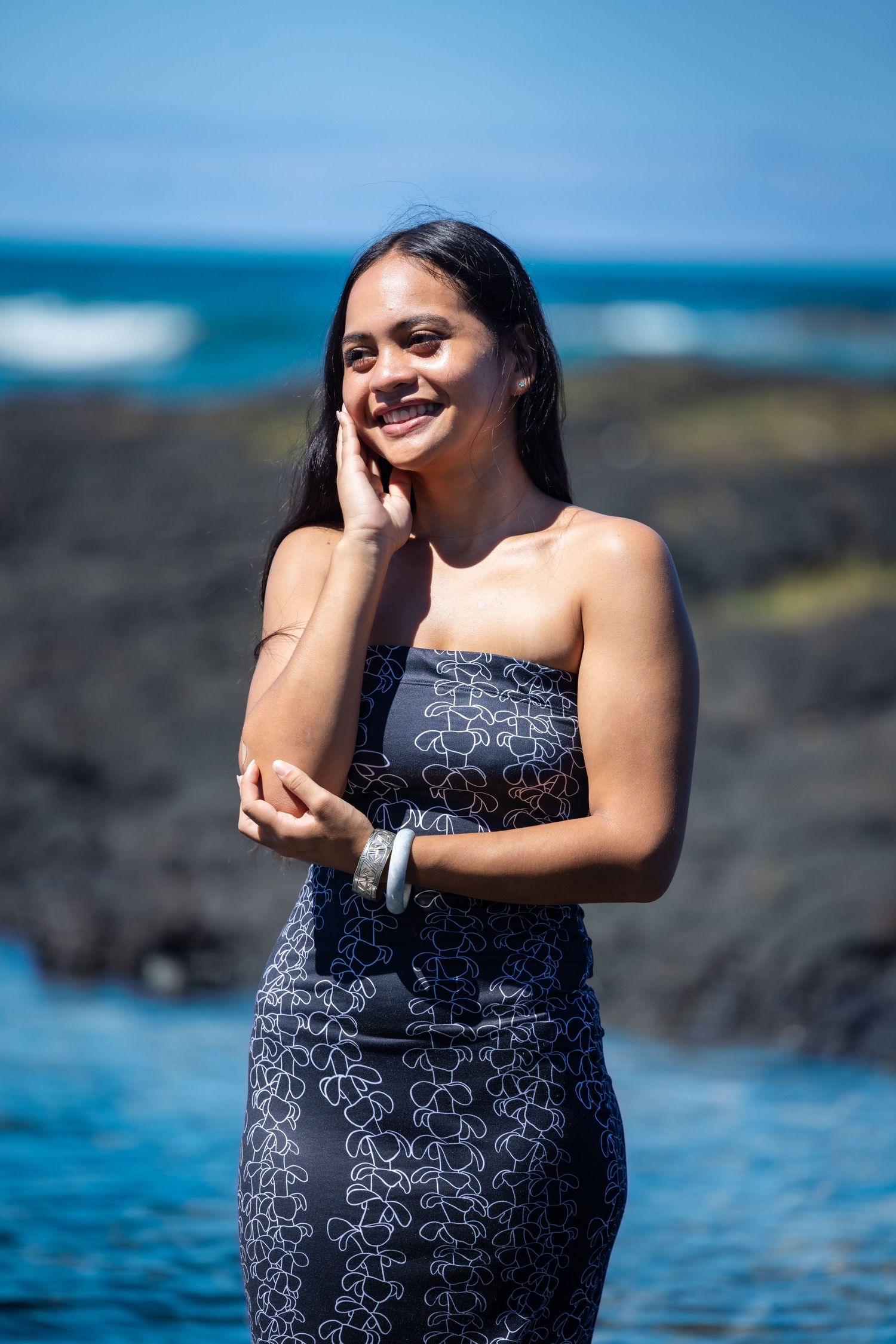 Woman in floral dress poses by rocky coastline with blue ocean waves.