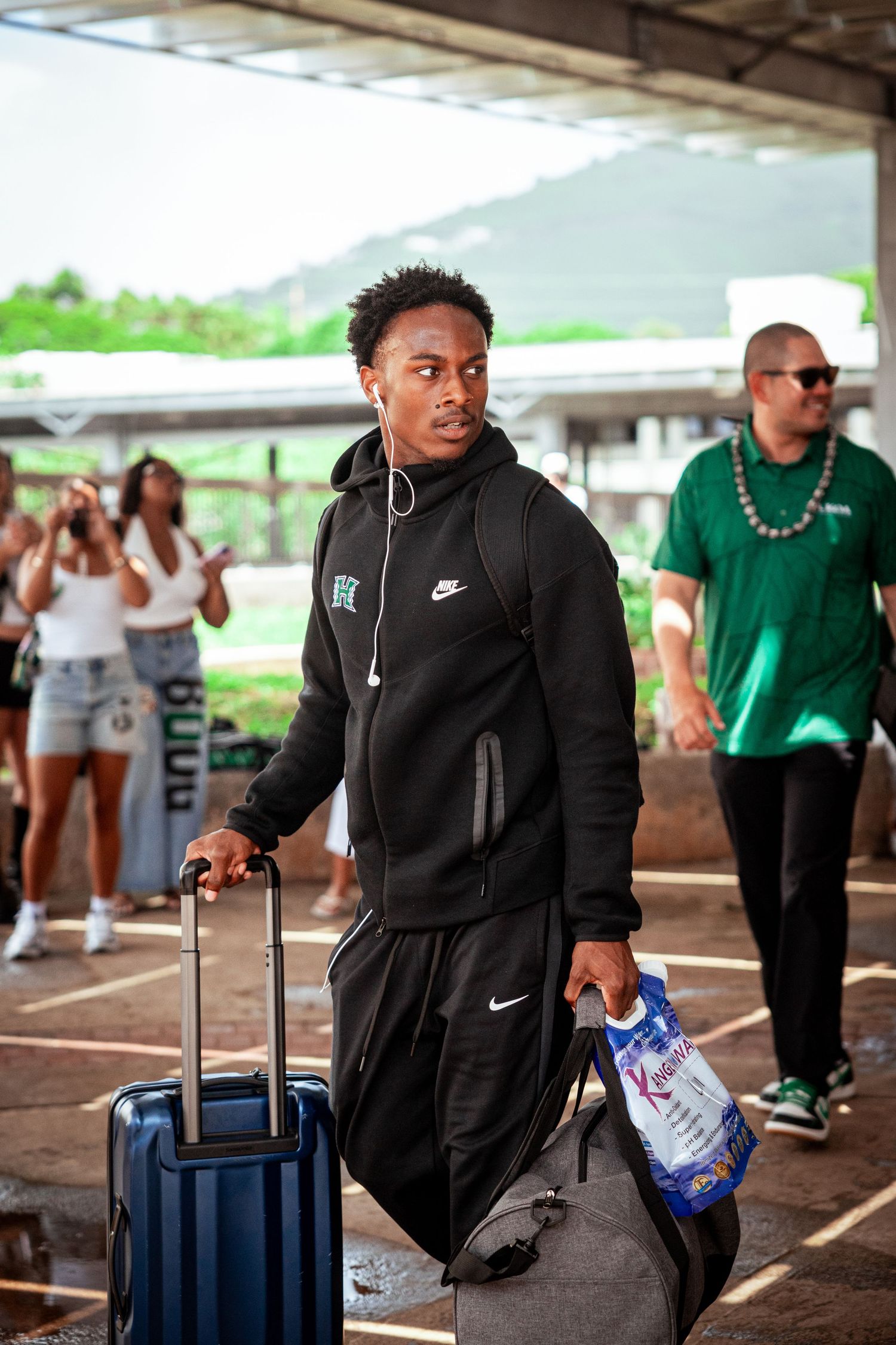 A young man in a black Nike hoodie pulls a blue suitcase while carrying a water bottle at an outdoor arrival area.