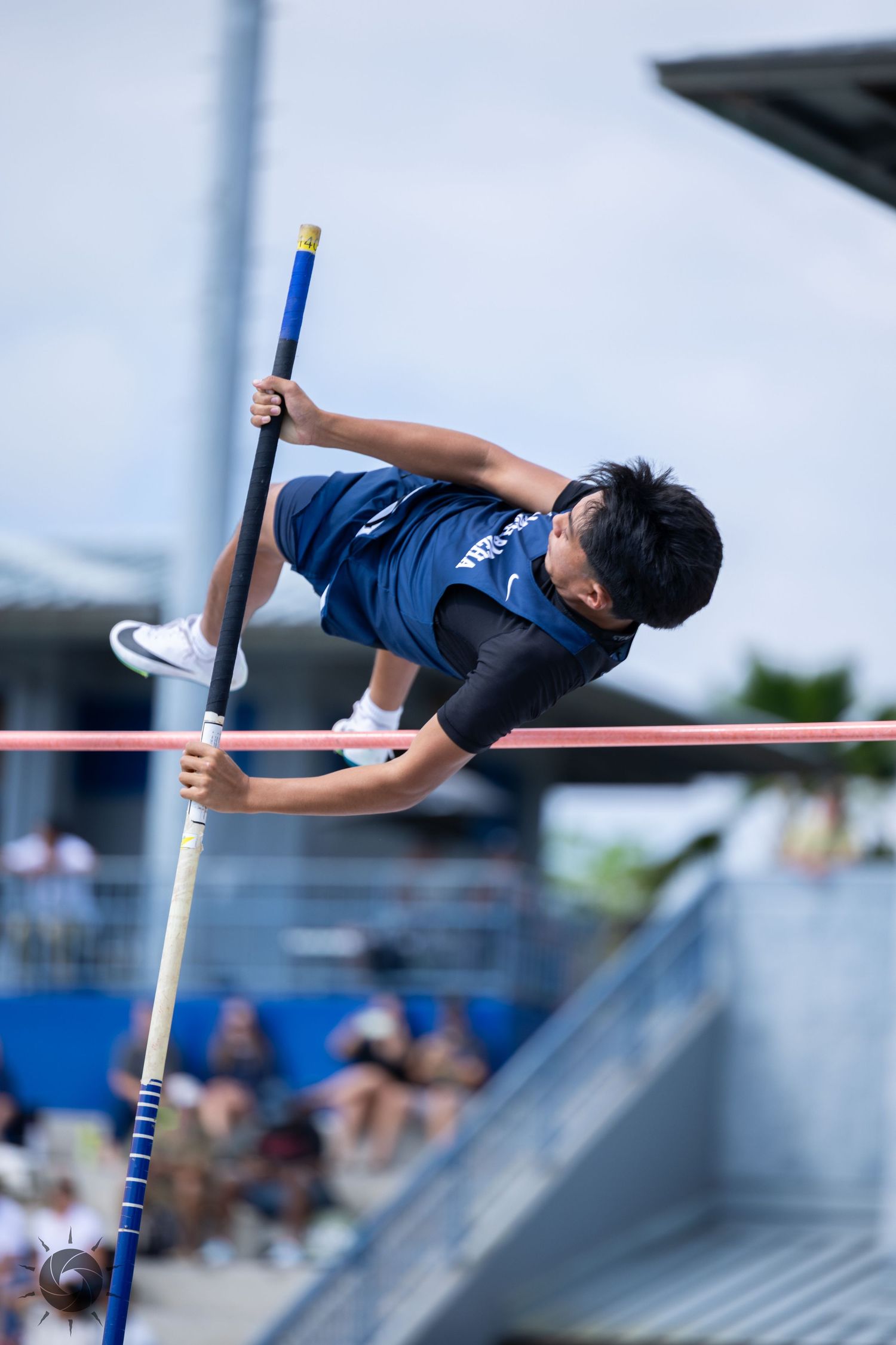High school athlete performs pole vault technique at track and field competition, arching over the bar in navy uniform.