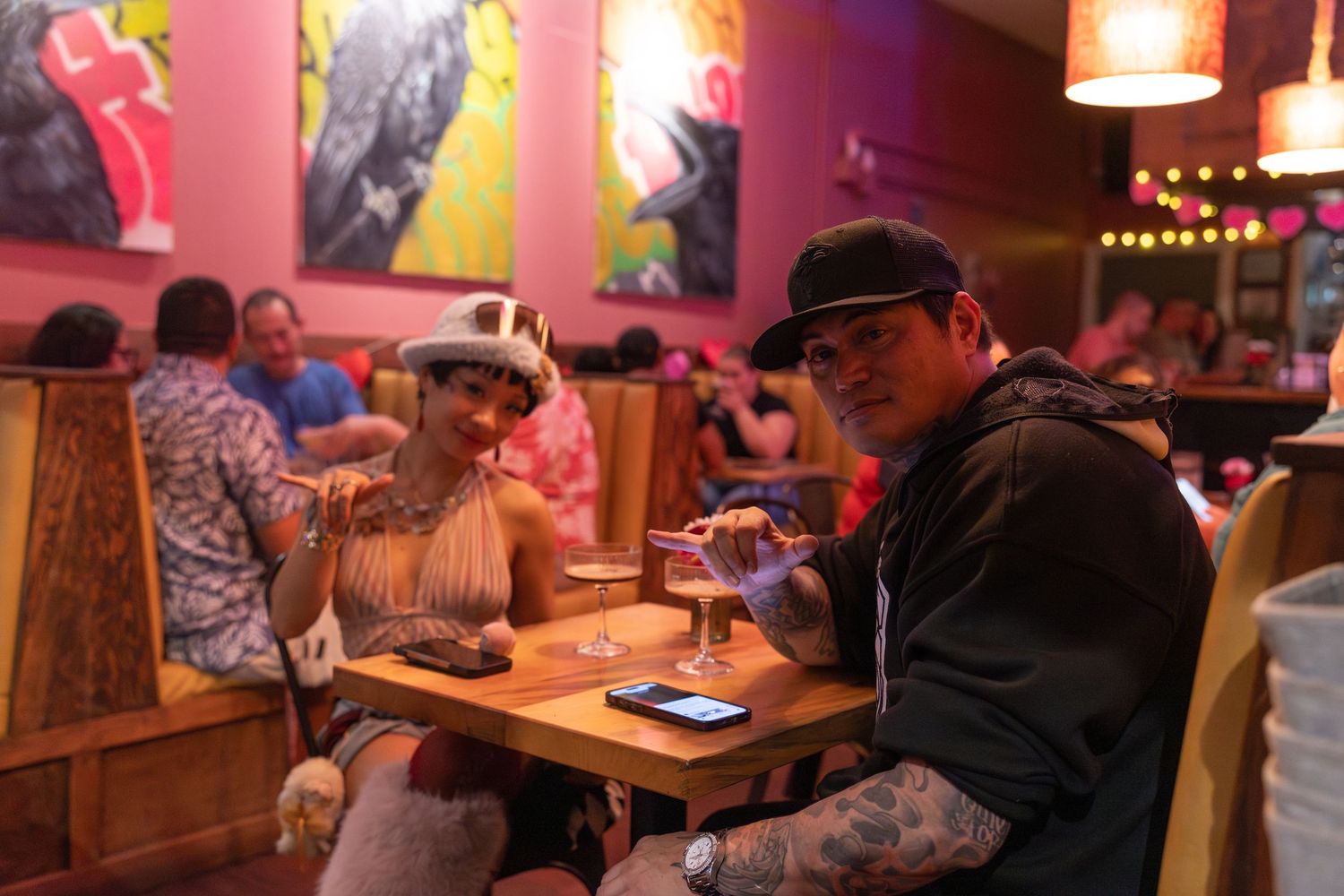 Patrons enjoy drinks and conversation in a dimly lit bar with warm pink lighting and wooden tables.