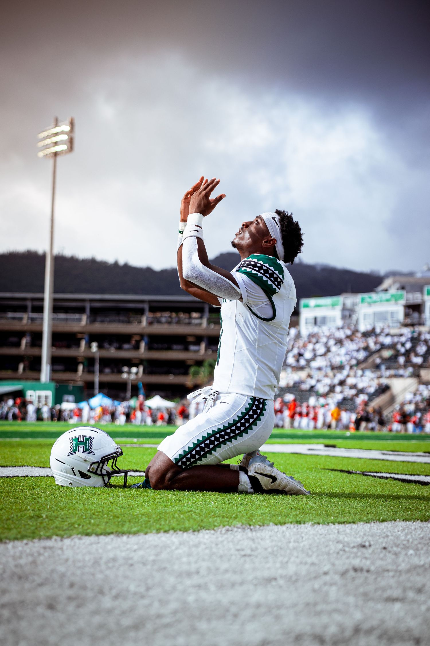 Hawaii Warriors football player kneels on field, arms raised skyward in celebration, wearing white uniform under dramatic cloudy sky.