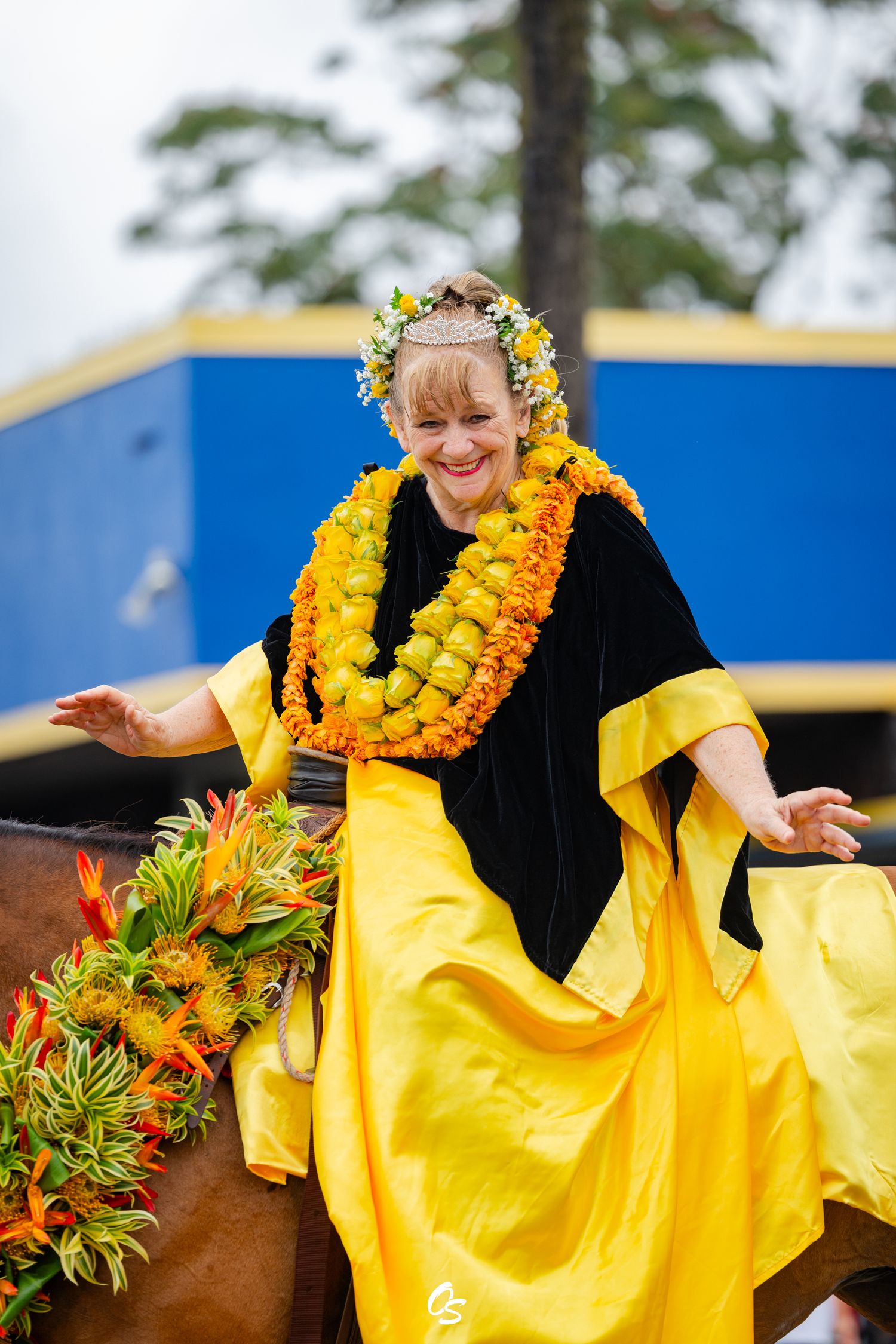 Woman in yellow dress and floral lei rides horse in Hawaiian parade celebration.